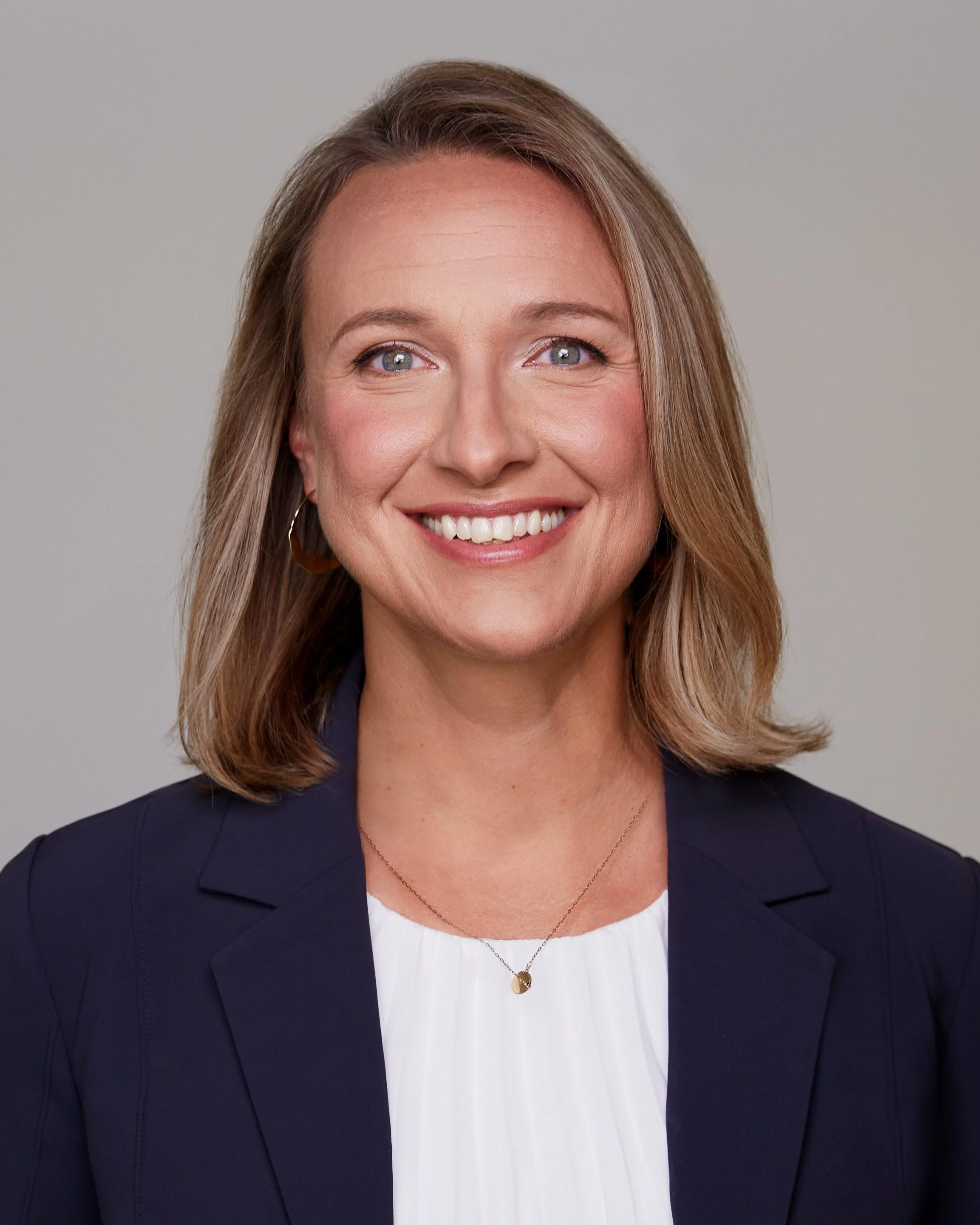A woman with shoulder-length blonde hair, wearing a navy blazer over a white top, a gold pendant necklace, and gold hoop earrings, smiling against a light gray background.