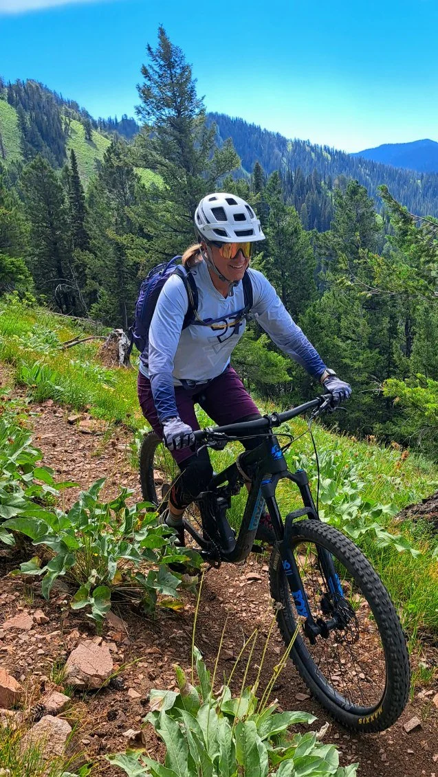 A woman mountain biking on a trail through a lush green forest with mountains in the background under a bright blue sky.