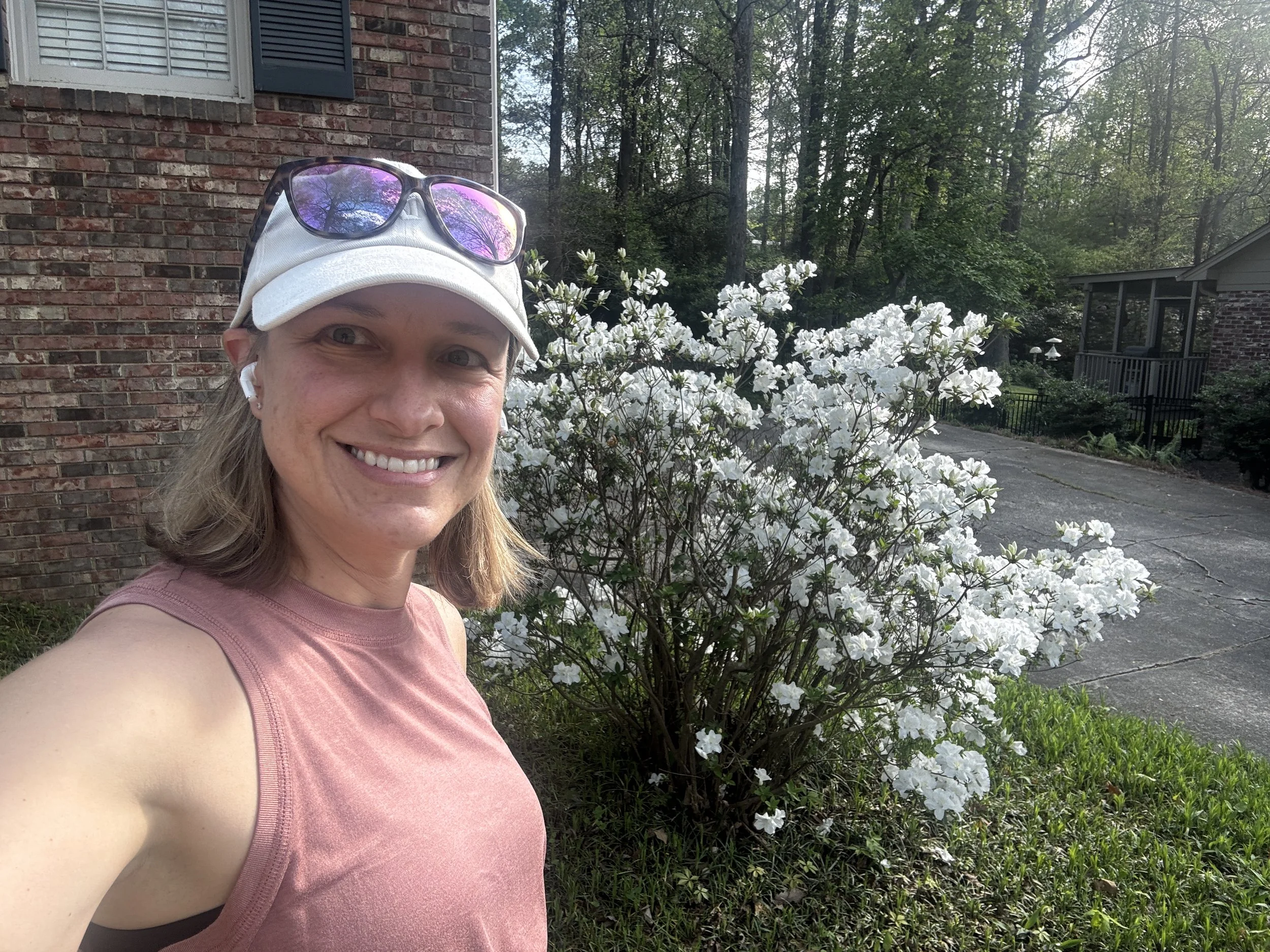 A smiling woman wearing a pink sleeveless top, white cap, and sunglasses on her cap, taking a selfie outdoors in front of a white flowering bush and a brick house with a window and another house in the background.