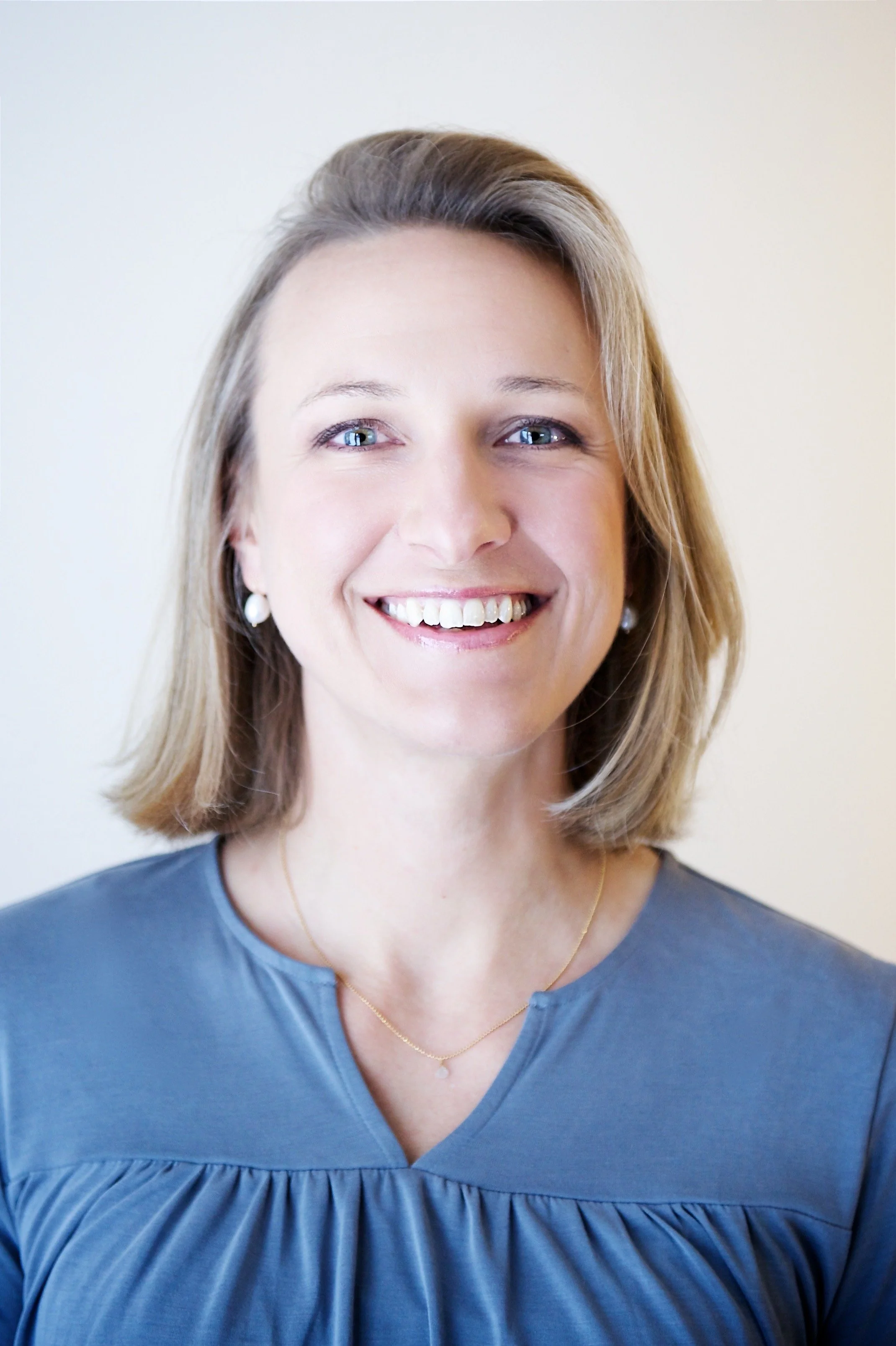 Close-up of a smiling woman with shoulder-length blonde hair, wearing a blue top and pearls against a plain background.