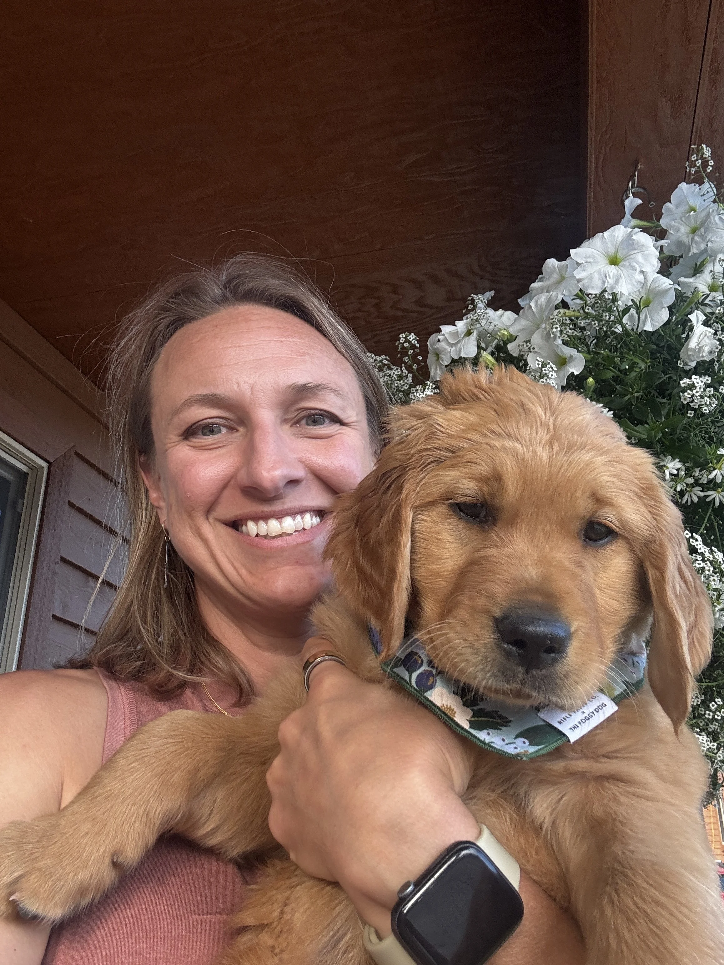 A woman smiling and holding a golden retriever puppy with a floral bandana, standing outside near a house with a wooden wall and white flowers in the background.