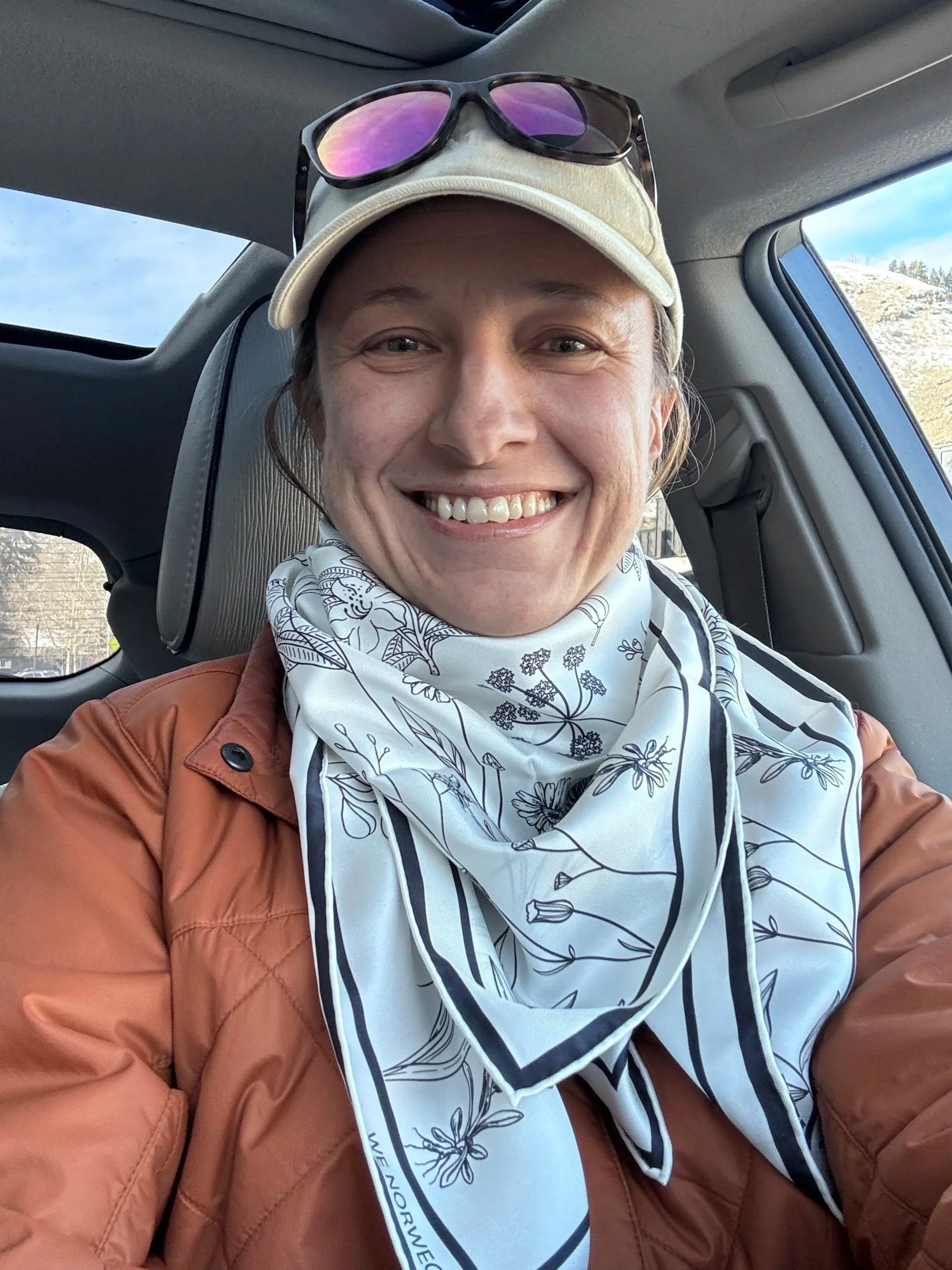 A smiling person wearing a beige cap with sunglasses on top, a white scarf with black floral patterns, and a brown jacket, sitting inside a vehicle with a window showing a landscape outside.
