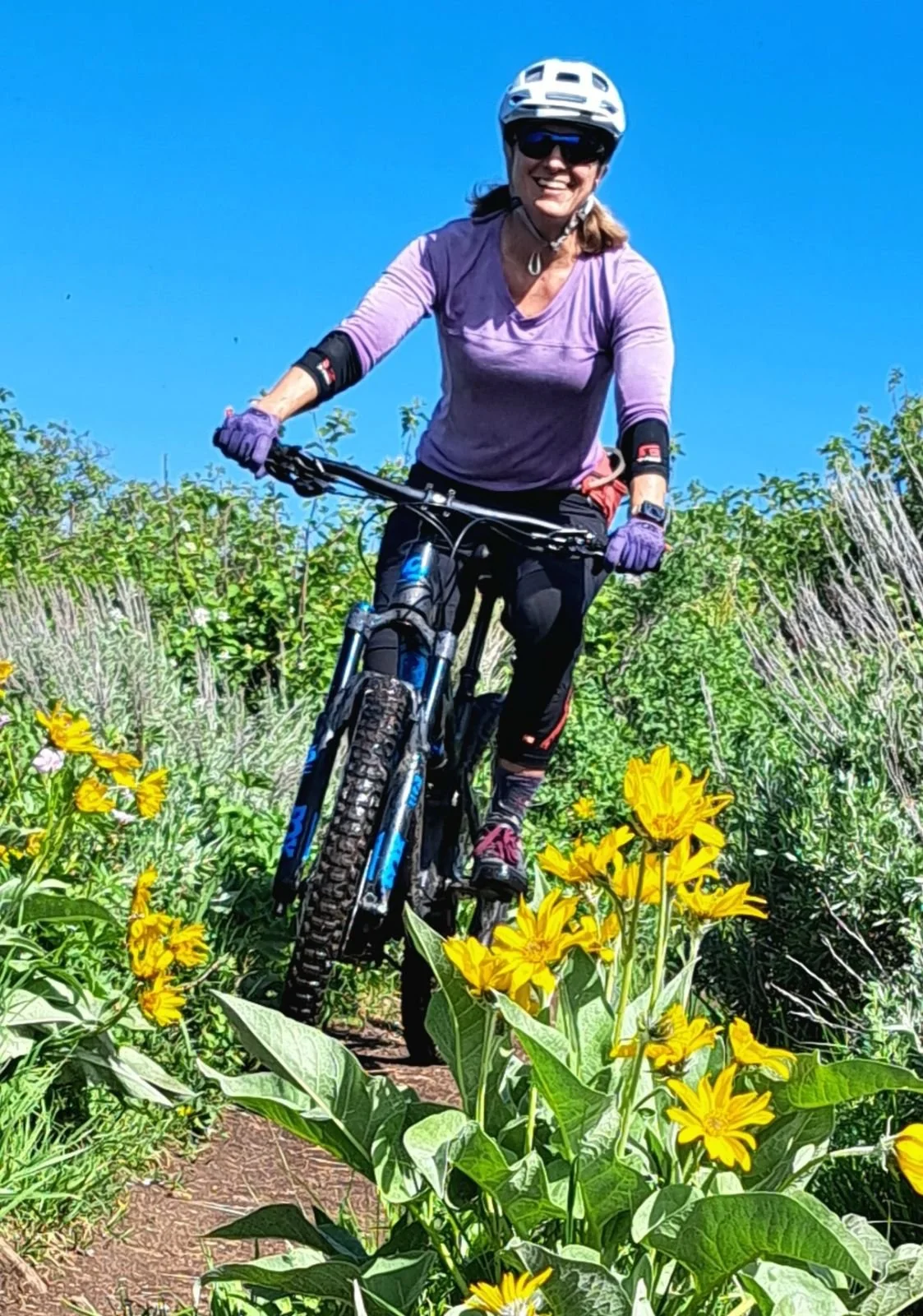 A woman mountain biking on a narrow trail through a field of yellow flowers, wearing a purple shirt, helmet, sunglasses, and gloves against a clear blue sky.