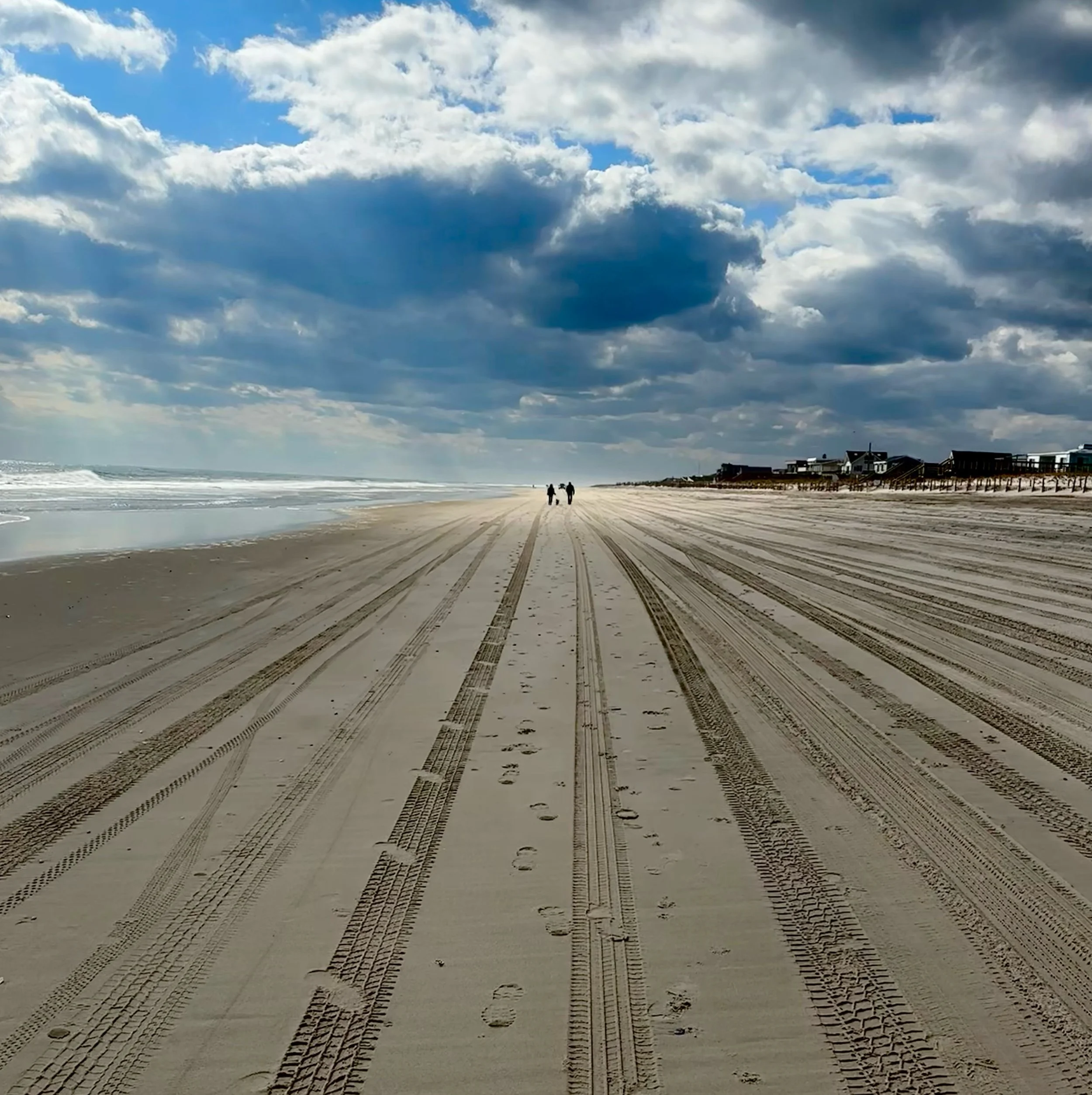 A sandy beach with tire tracks and footprints, two people walking in the distance, ocean waves on the left, and a partly cloudy sky overhead, with beach houses along the horizon.