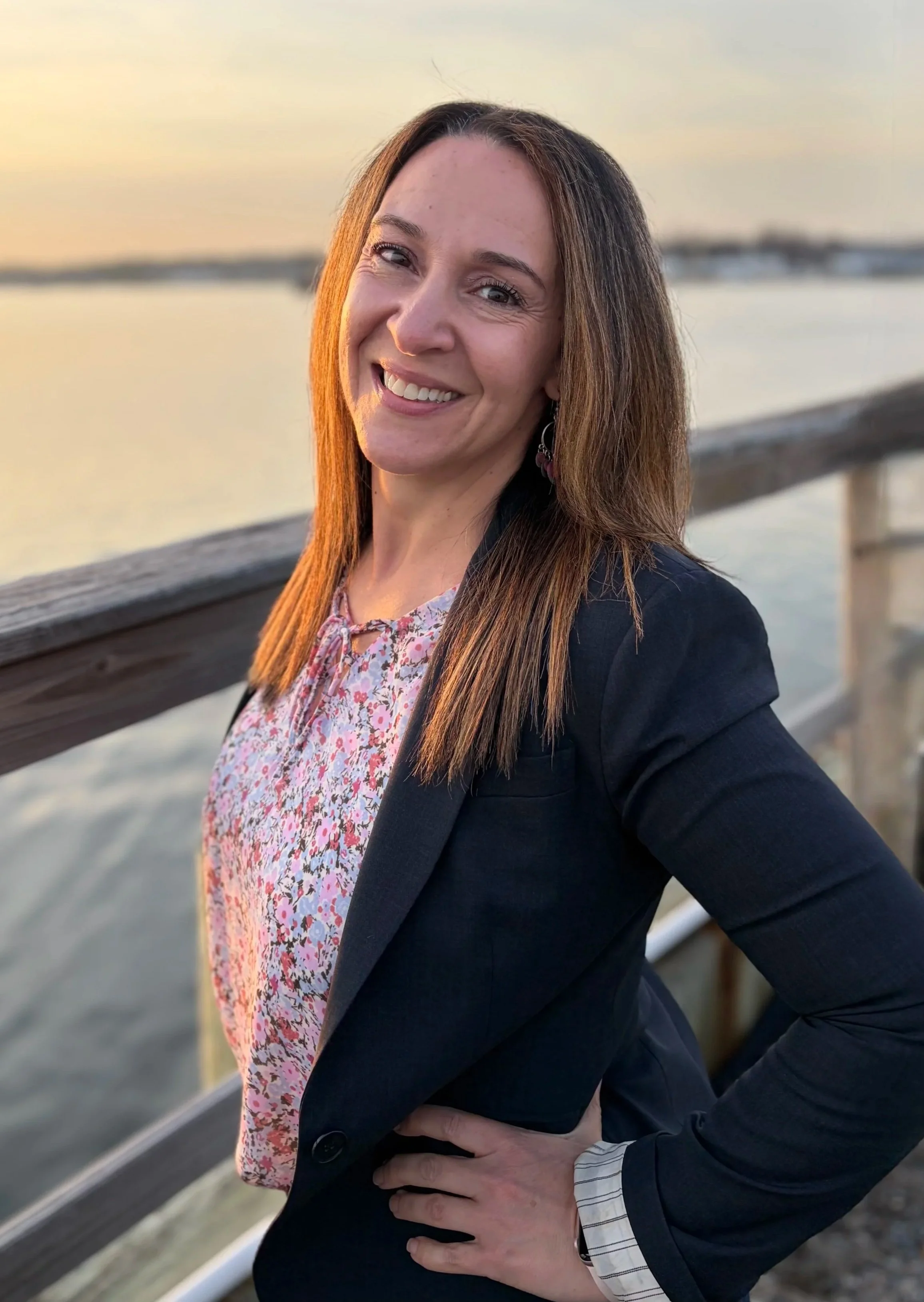 A woman with long brown hair smiling at the camera, standing outdoors on a wooden dock with water and a sunset background.