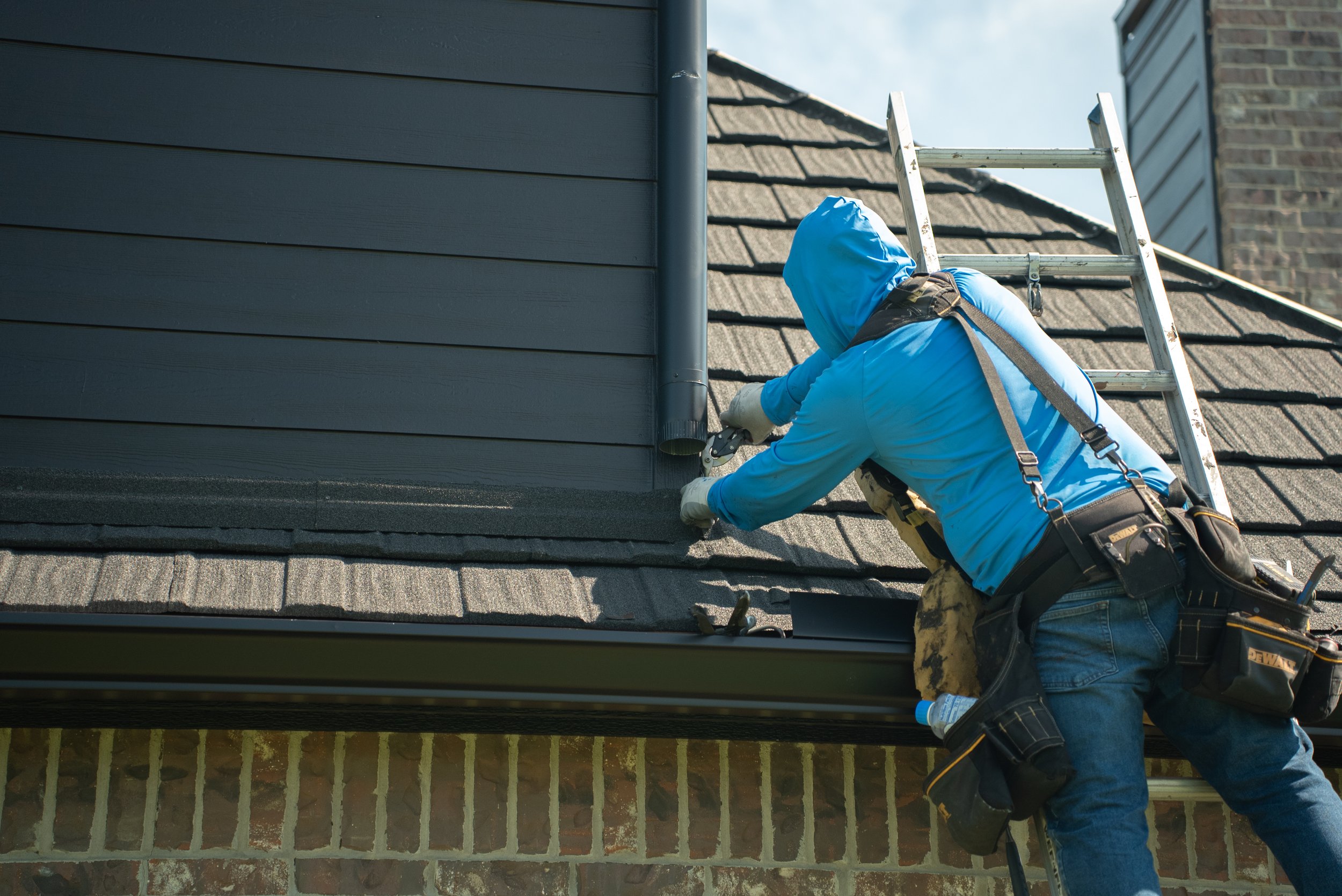 A worker wearing a blue hoodie, gloves, and tool belt, installing a black drain pipe on the roof of a house, with a ladder leaning against the roof.