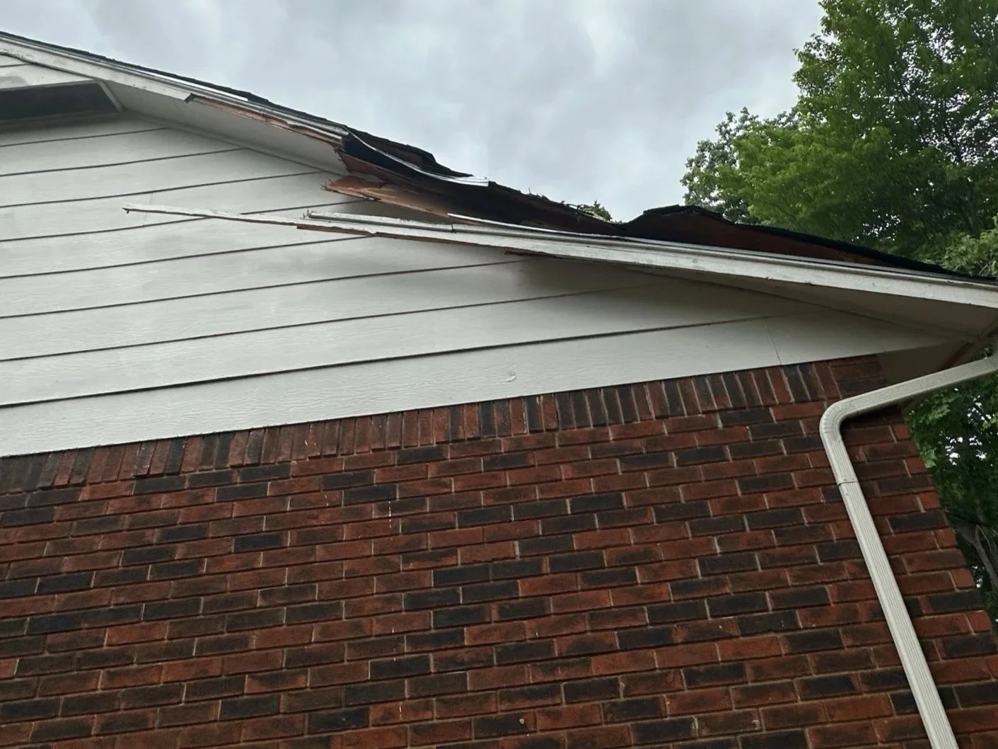 Part of a house showing the roof with damaged shingles and the exterior wall made of red brick, also a white gutter on the side.