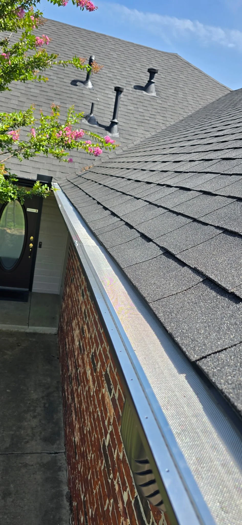 Close-up view of a shingled roof with four roof vents, part of a brick wall, and a tree with pink flowers in the foreground, under a blue sky.