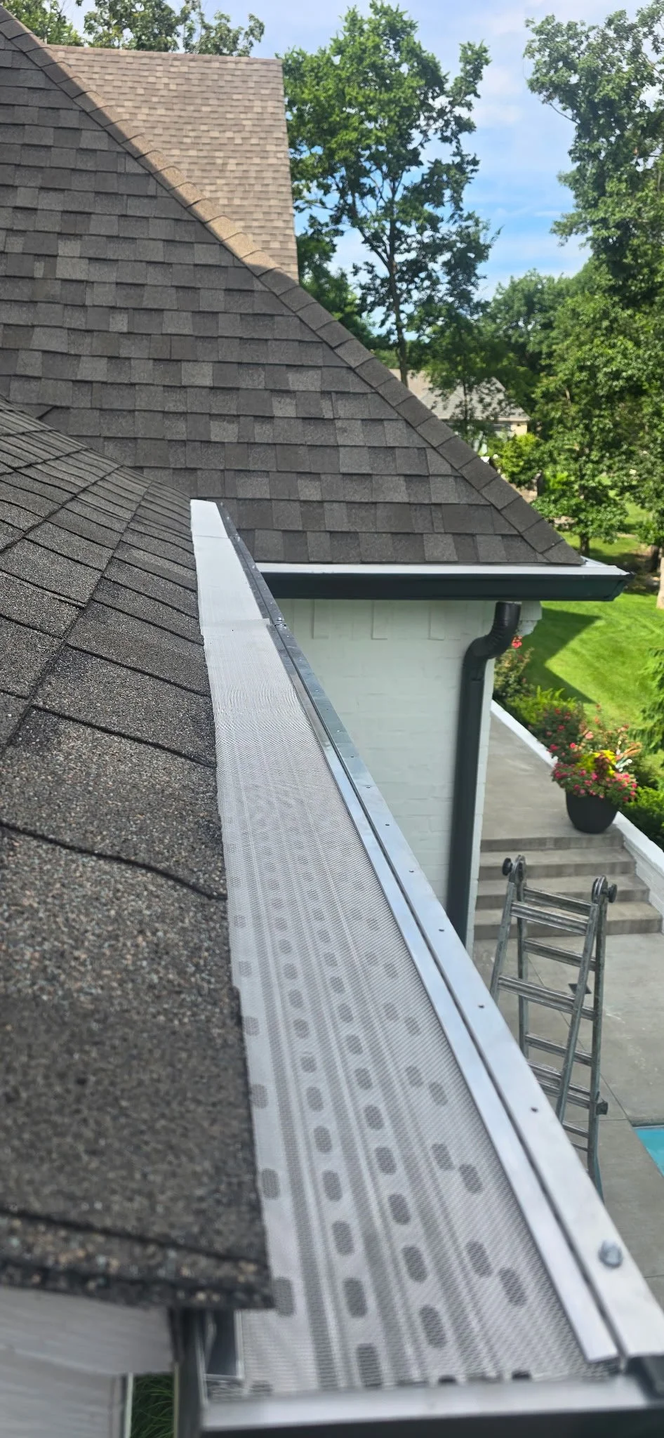 Close-up of a roof with asphalt shingles, showing a metal flashing along the edge and a ladder on the ground nearby.