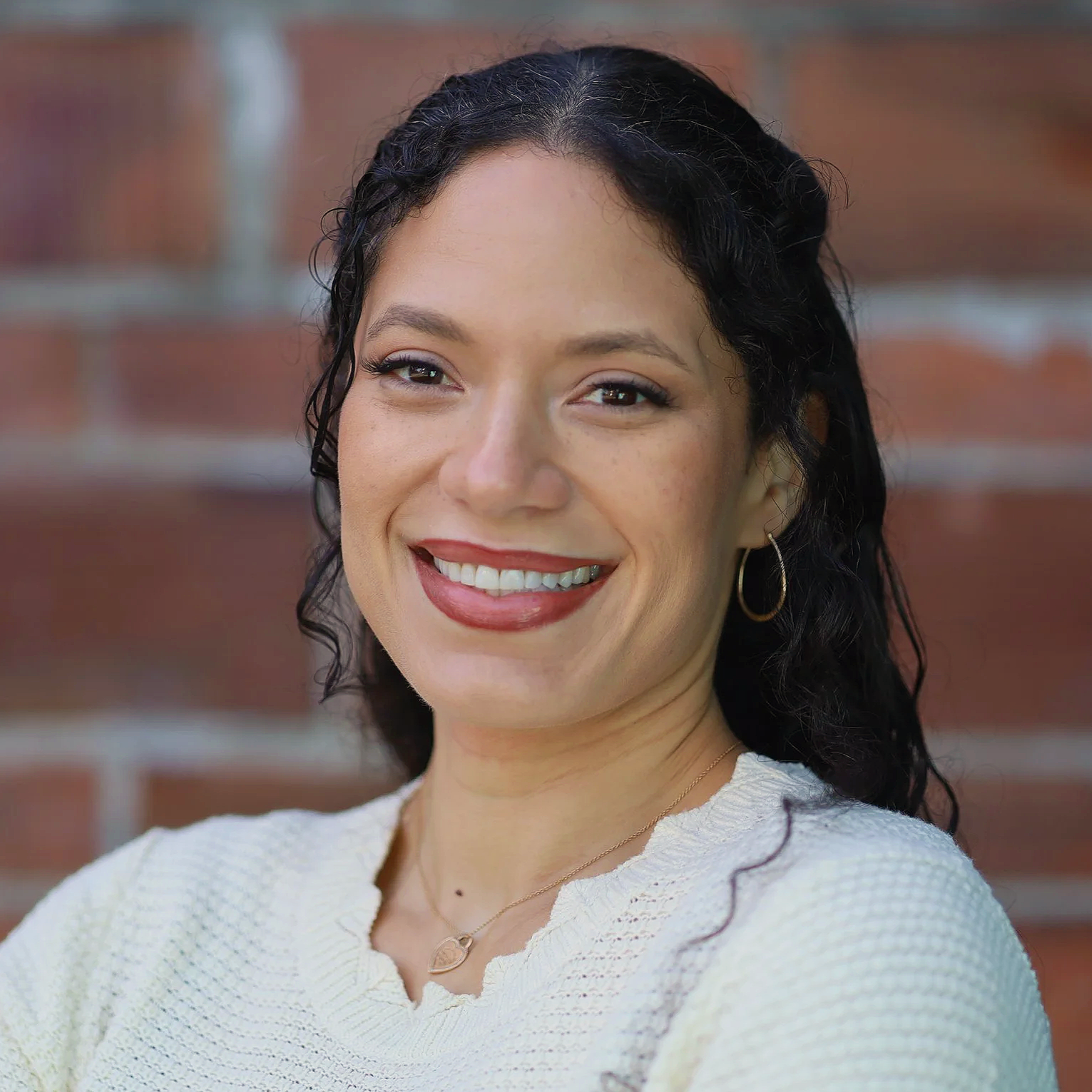 Close-up portrait of a woman smiling, with dark curly hair, wearing gold hoop earrings, a gold necklace with a heart pendant, and a creamtextured top, standing against a brick wall background.