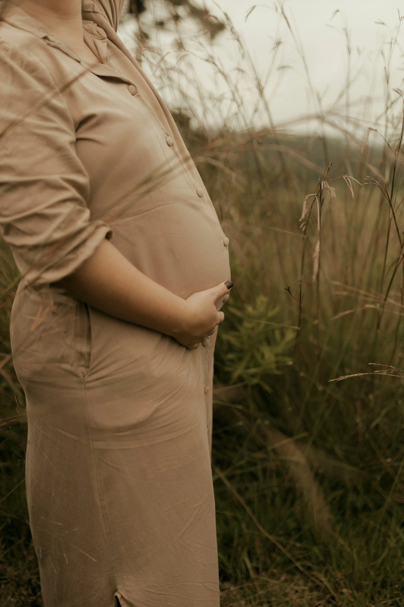 A pregnant woman holding her belly with both hands, standing in a field of tall grass during sunset.