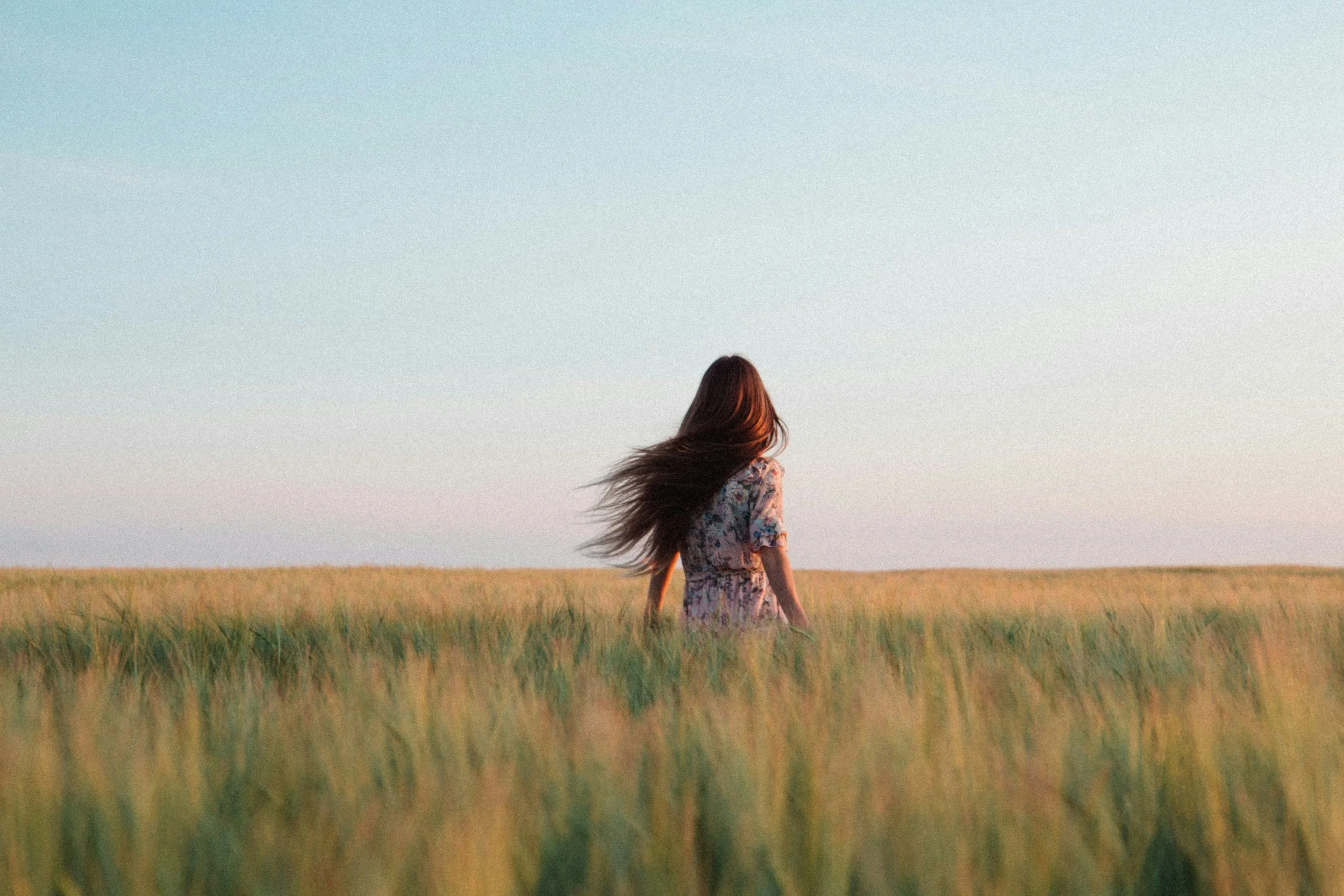A woman with long hair wearing a floral dress sitting in a field of green grass, facing away, with a clear sky in the background.