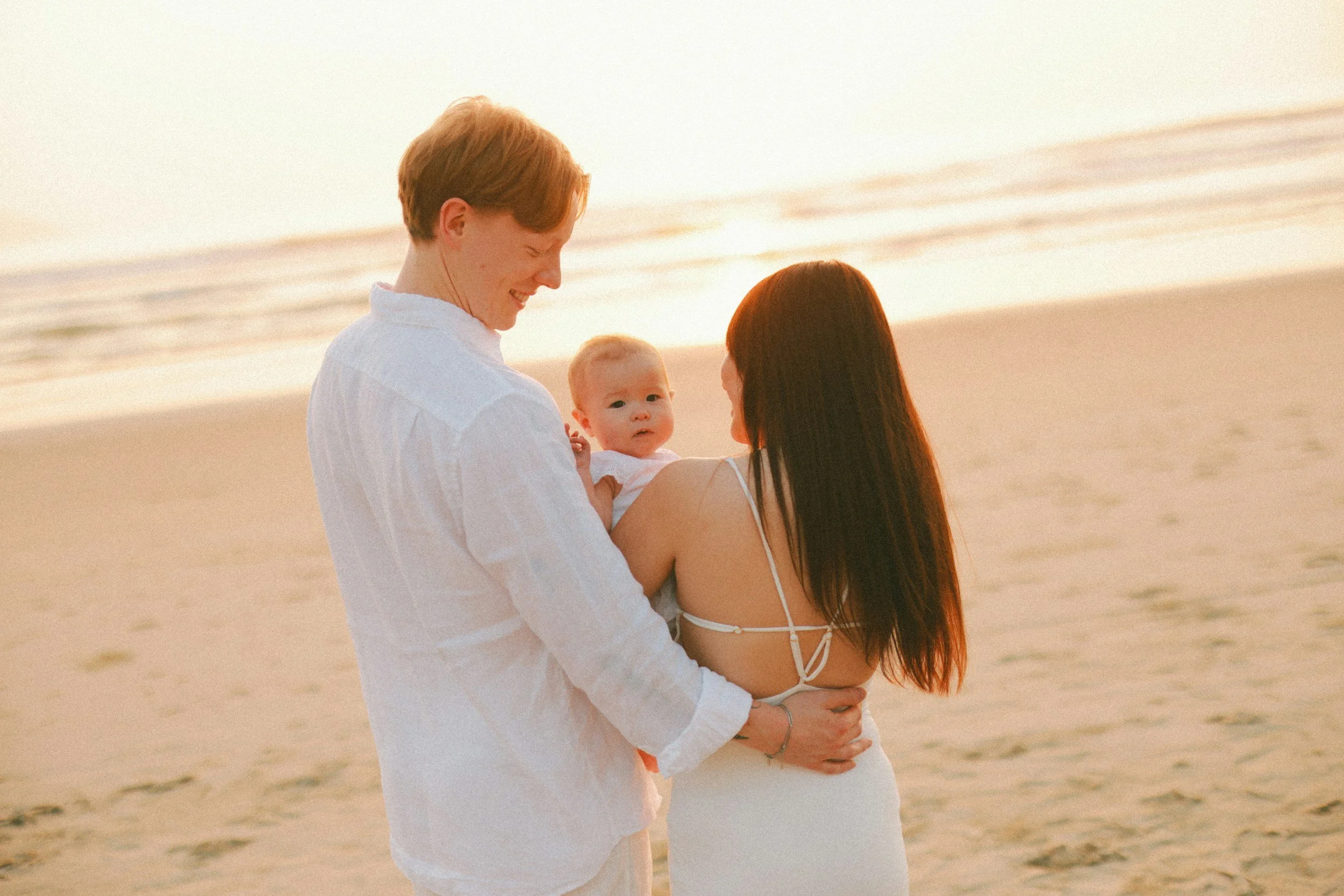 A family at the beach during sunset, with a man, woman, and baby enjoying the moment.