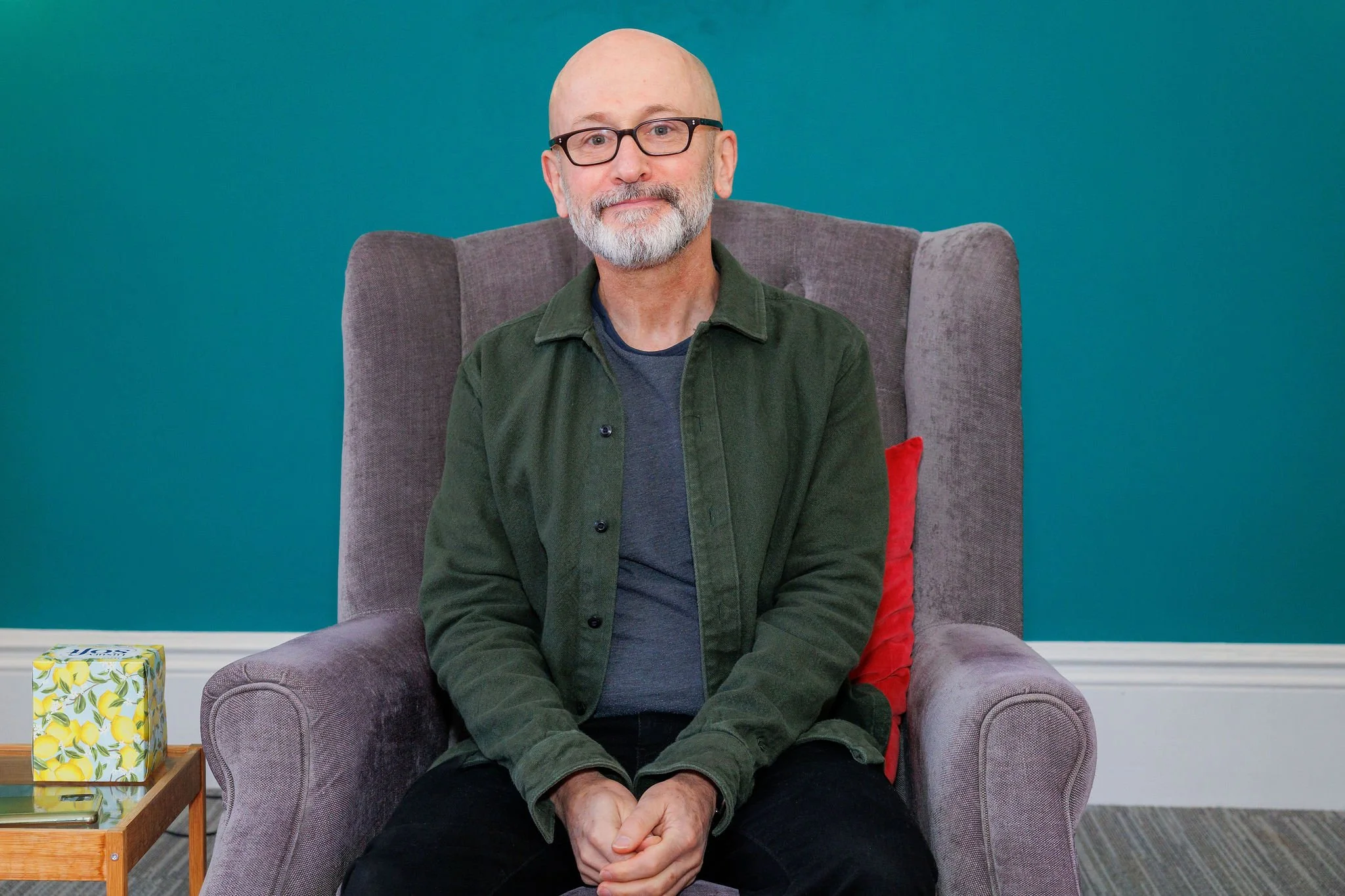 Aidan Harvey with a beard and glasses sitting in a grey armchair against a teal wall. He is wearing a green jacket over a dark shirt and has his hands clasped in his lap.