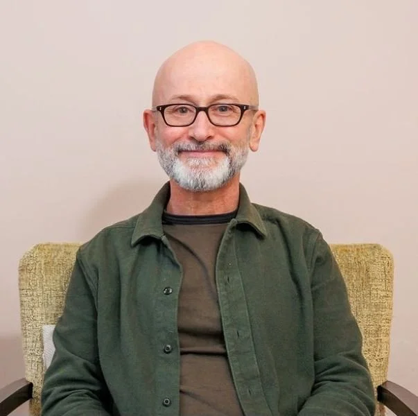 Aidan Harvey with glasses and a beard, sitting on a greenish-yellow chair against a plain beige wall, smiling at the camera.
