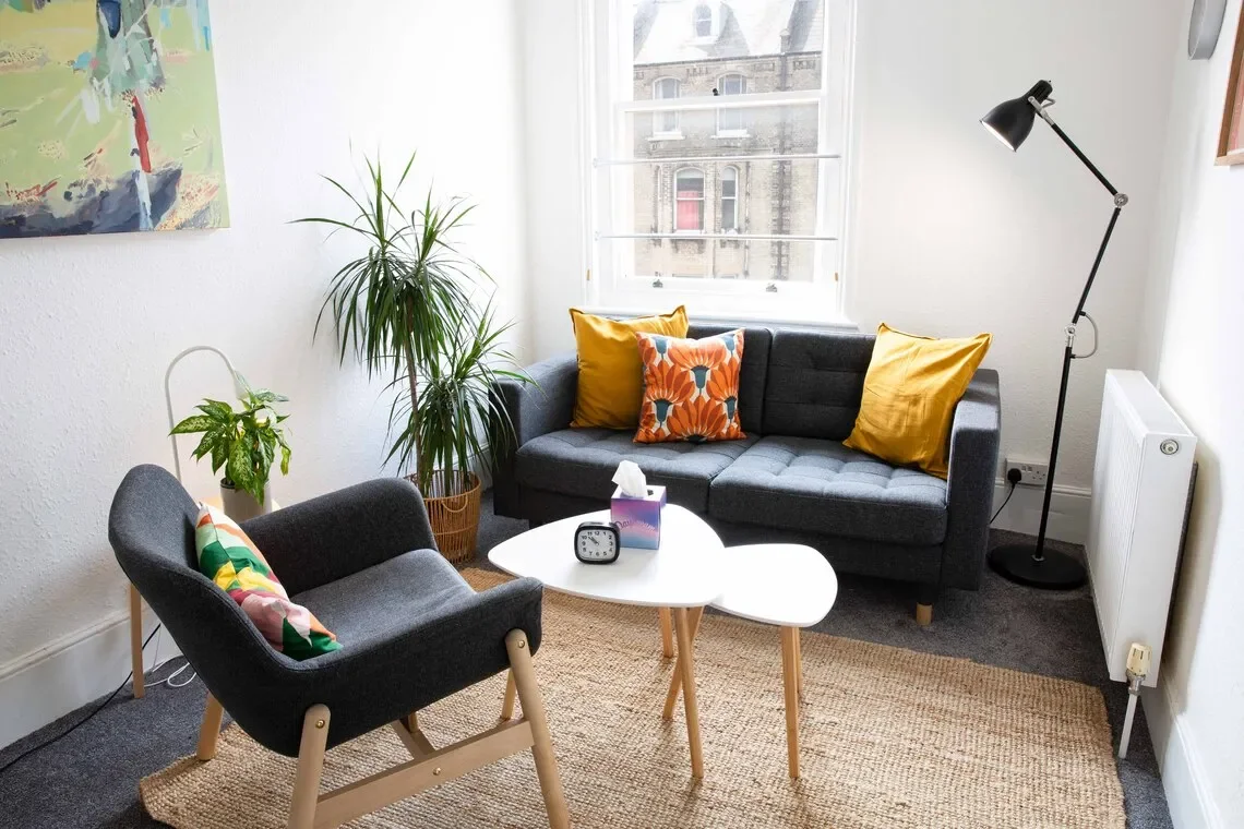 A cozy room at Murmuration Therapy Rooms - with a dark gray sofa, a black armchair with wooden legs, colourful cushions, a white coffee table, potted plants, a tall window, and a colourful painting on the wall.