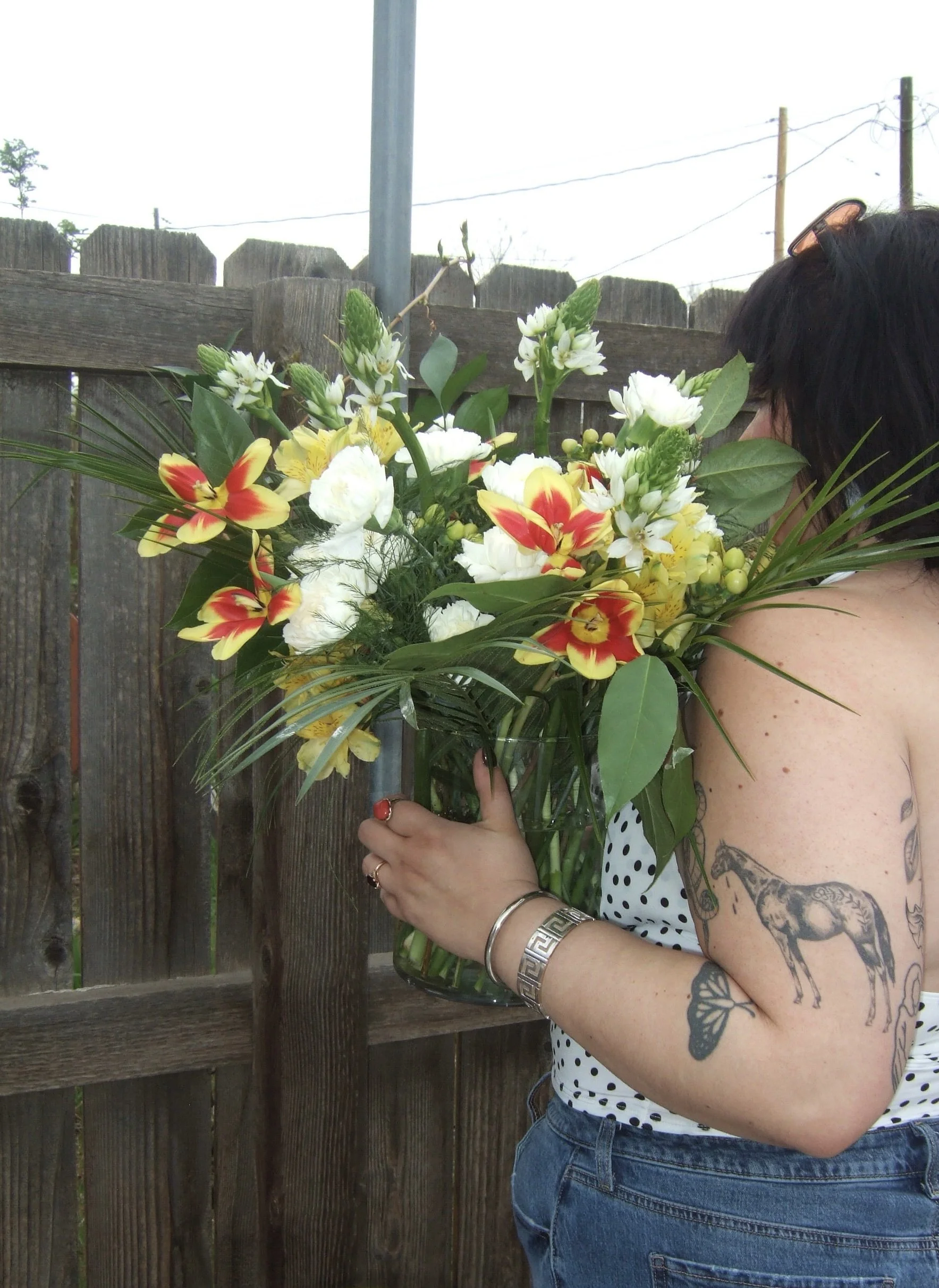 A woman holding a large bouquet of assorted flowers, including white, yellow, and red flowers, near a wooden fence outdoors.