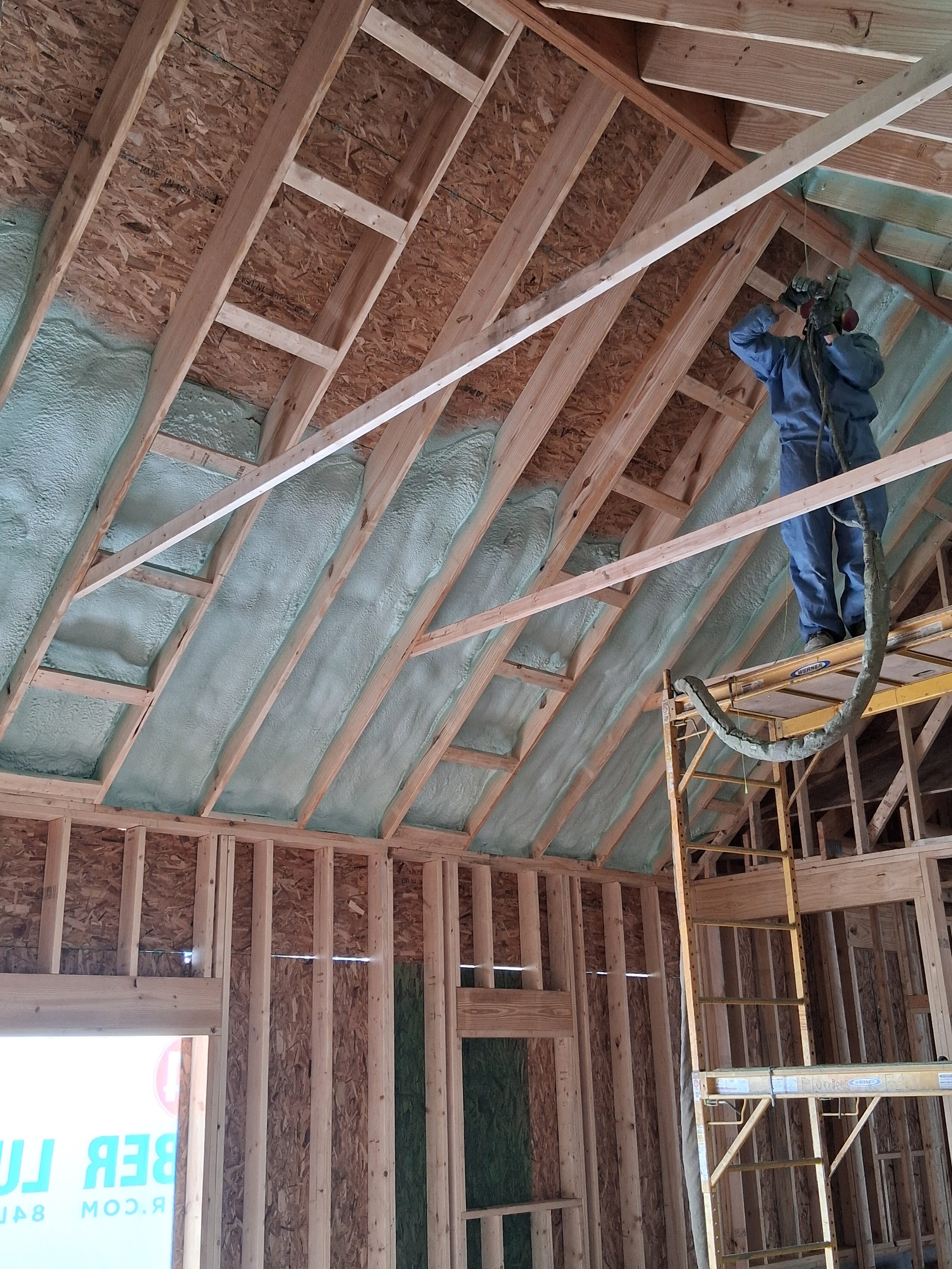 A construction worker in blue overalls standing on scaffolding, working on the wooden framing and insulation of a house's roof.