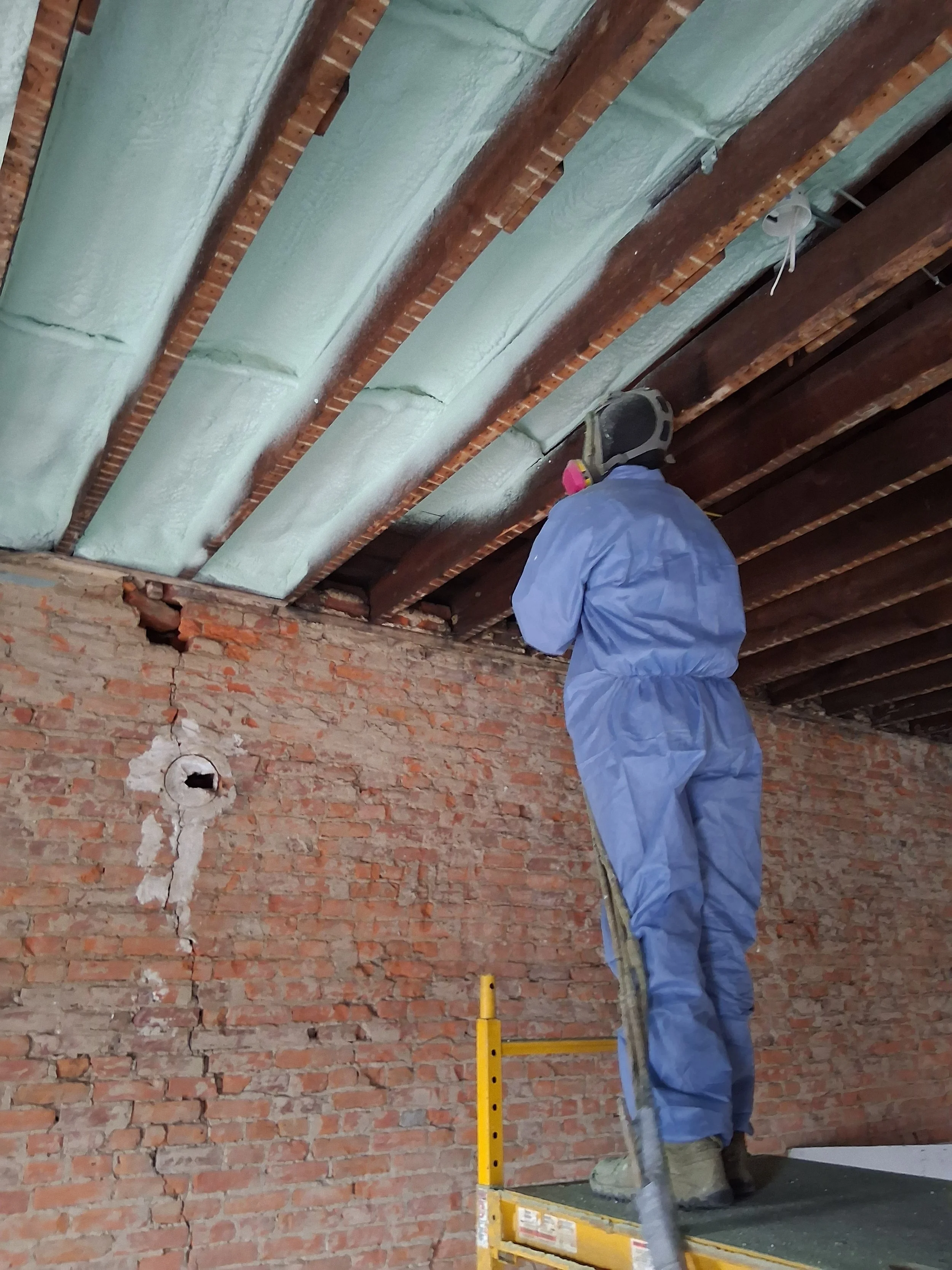 A worker in blue coveralls and a respirator is standing on a yellow scaffold, insulating a wooden ceiling with foam insulation.