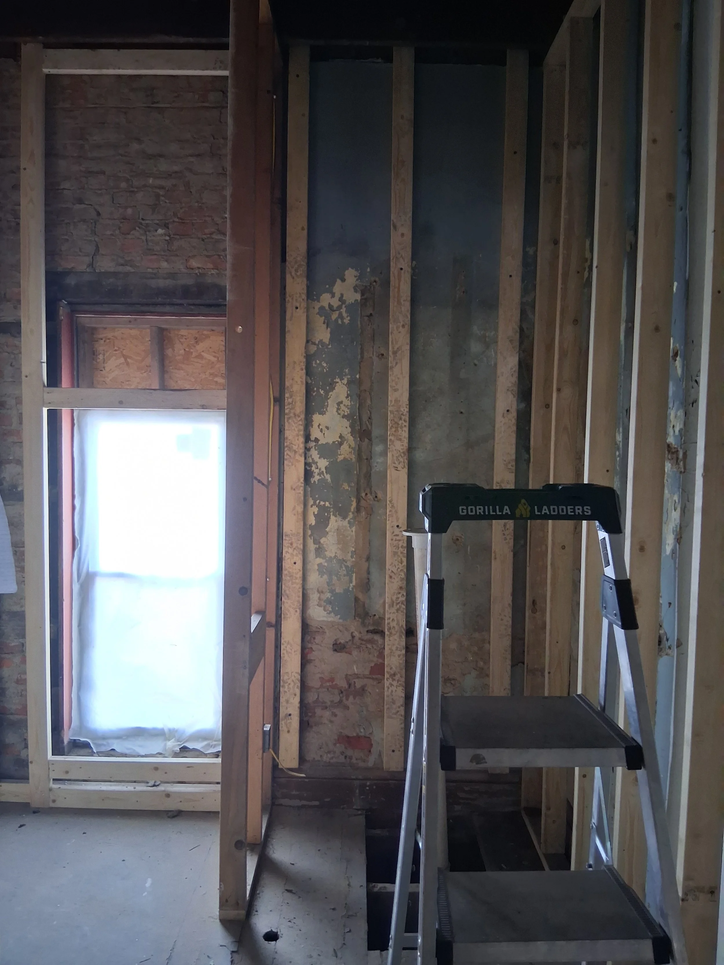 Room under construction with exposed wooden framing, a window with a white curtain, and a step ladder labeled 'Gorilla Ladders'.