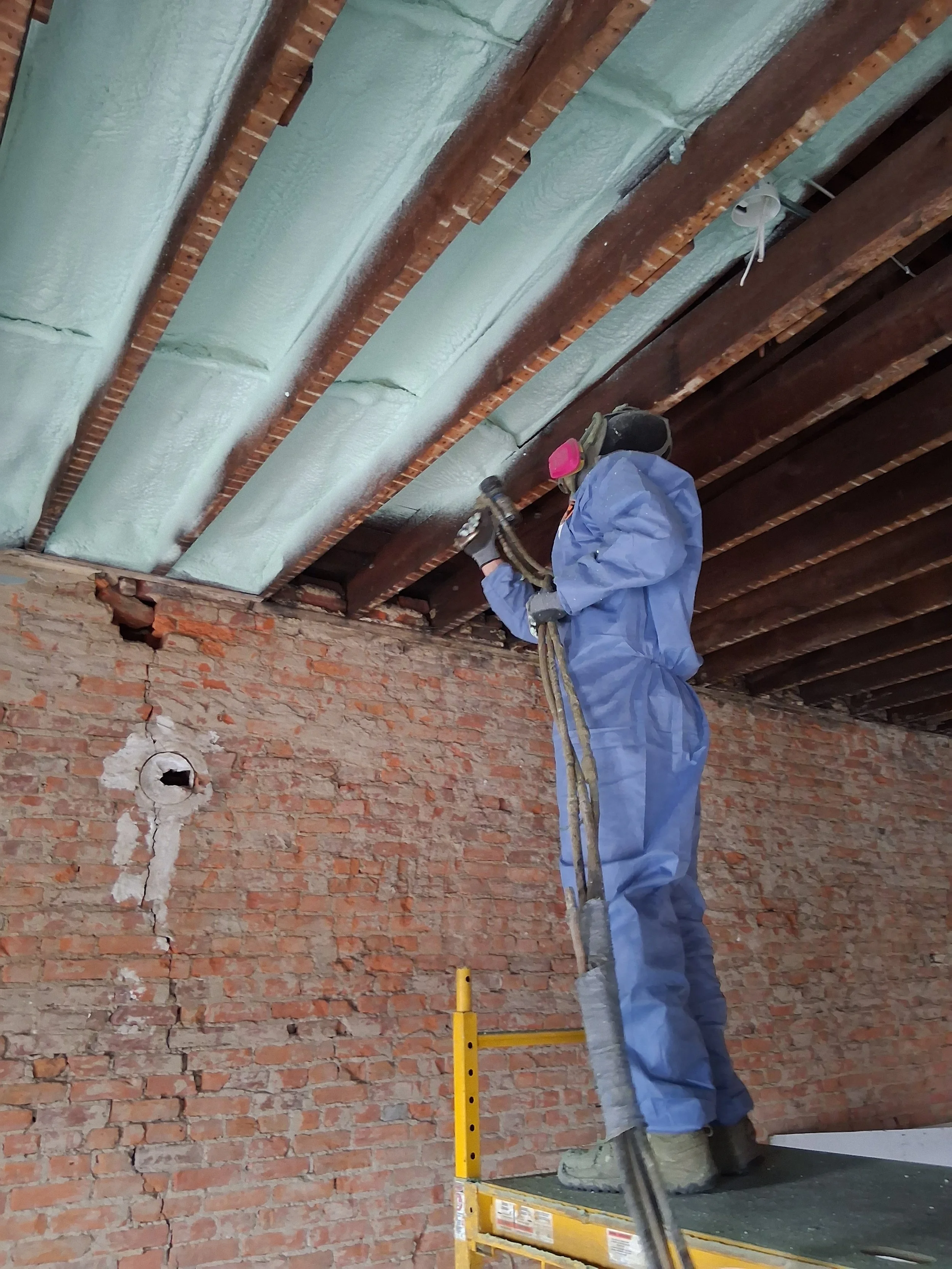 Construction worker in protective gear standing on a scaffold, spray painting or insulating a ceiling with silver insulation material in a building under renovation.