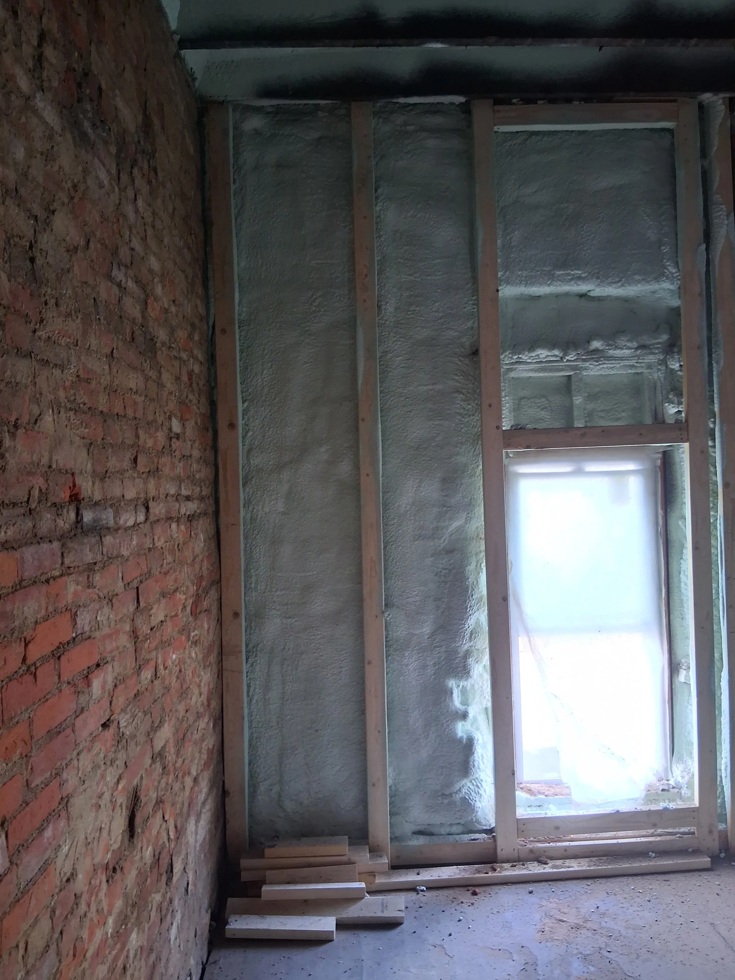 Interior of a room under renovation with exposed brick wall and wooden framing for a wall, with plastic sheeting on the window to block sunlight.
