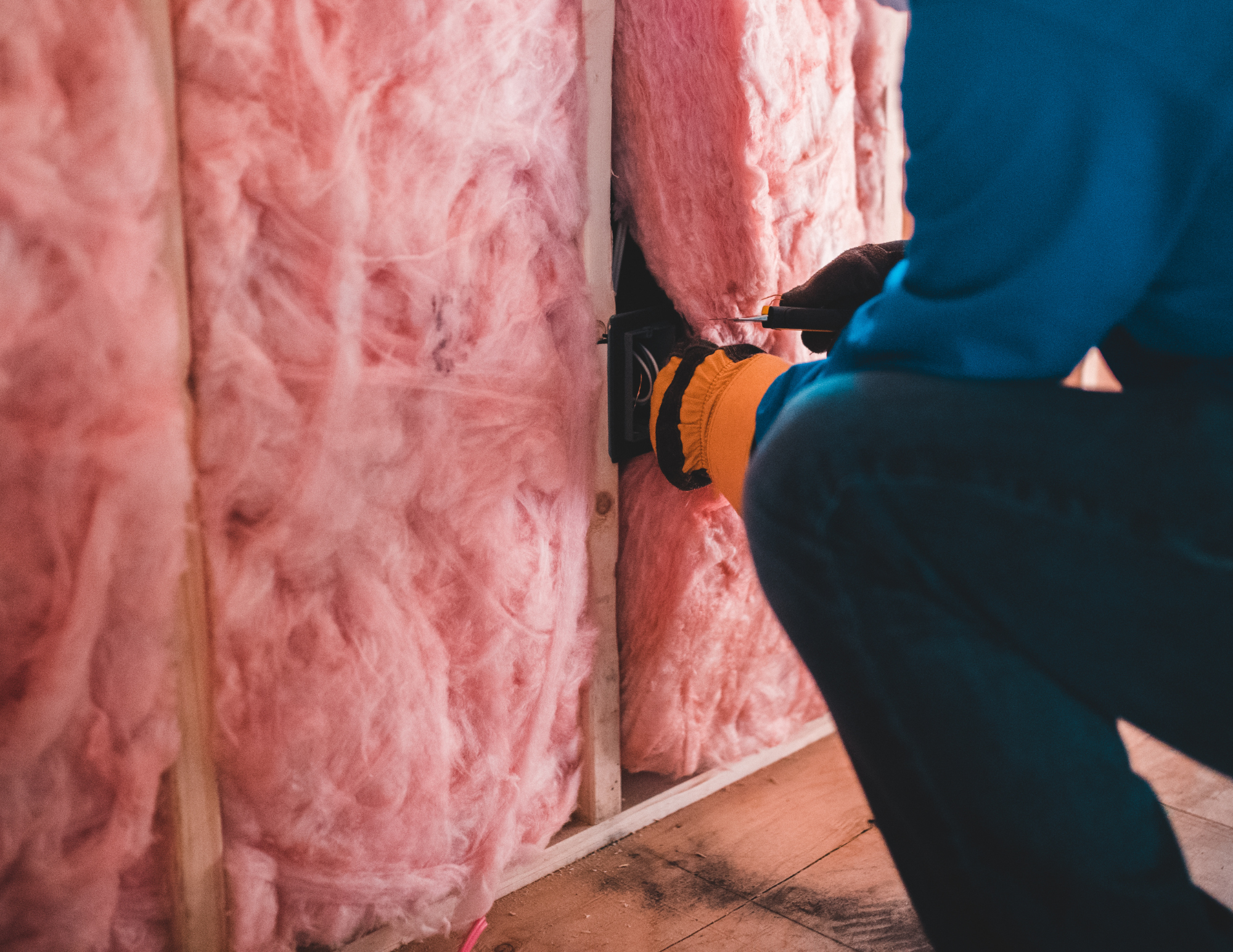 Person working on wall insulation, wearing gloves and blue jacket, using a screwdriver in a partially finished wall with pink fiberglass insulation.