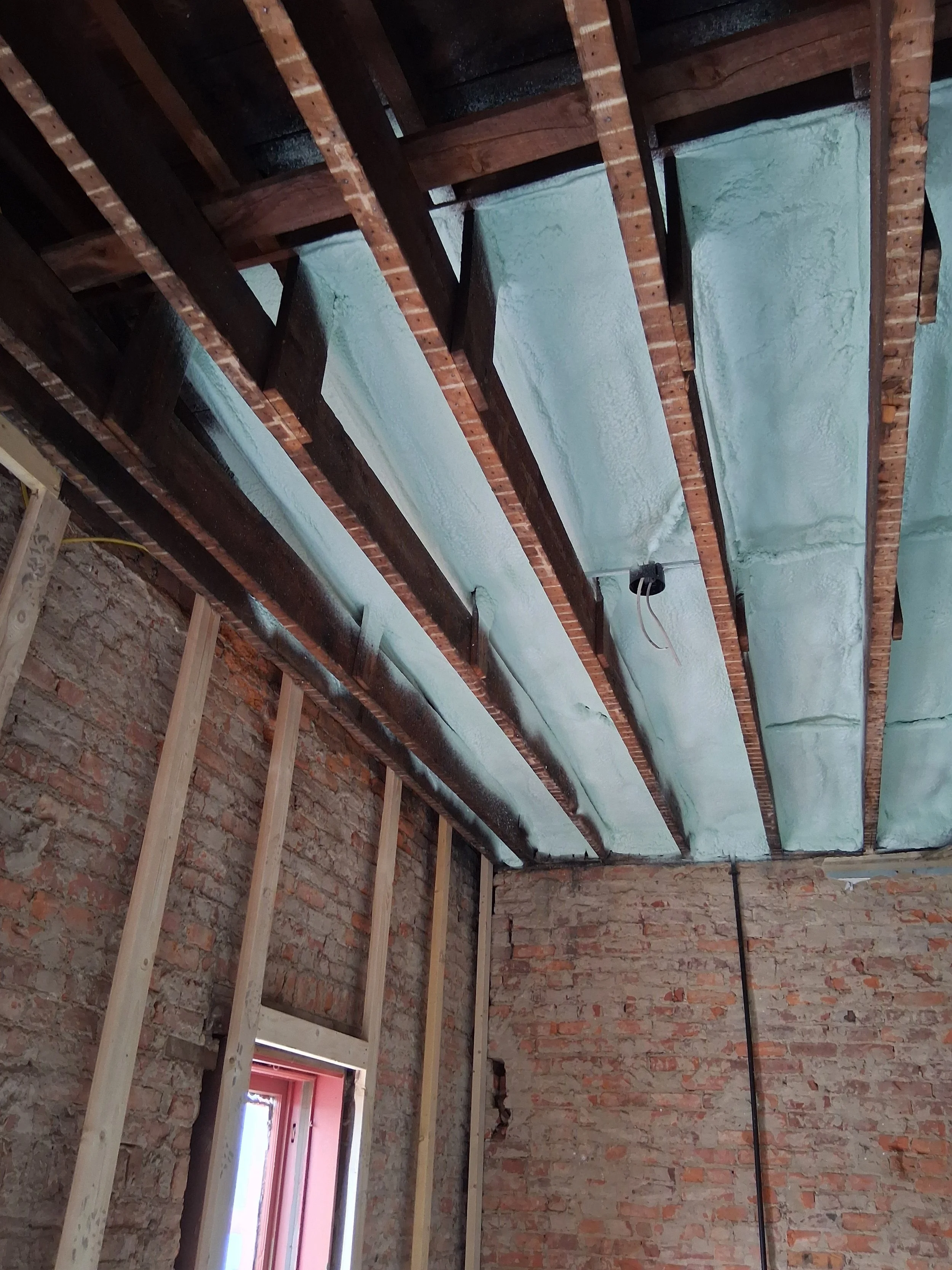 Interior of a room under construction with exposed brick walls, wooden framing, and spray foam insulation on the ceiling.
