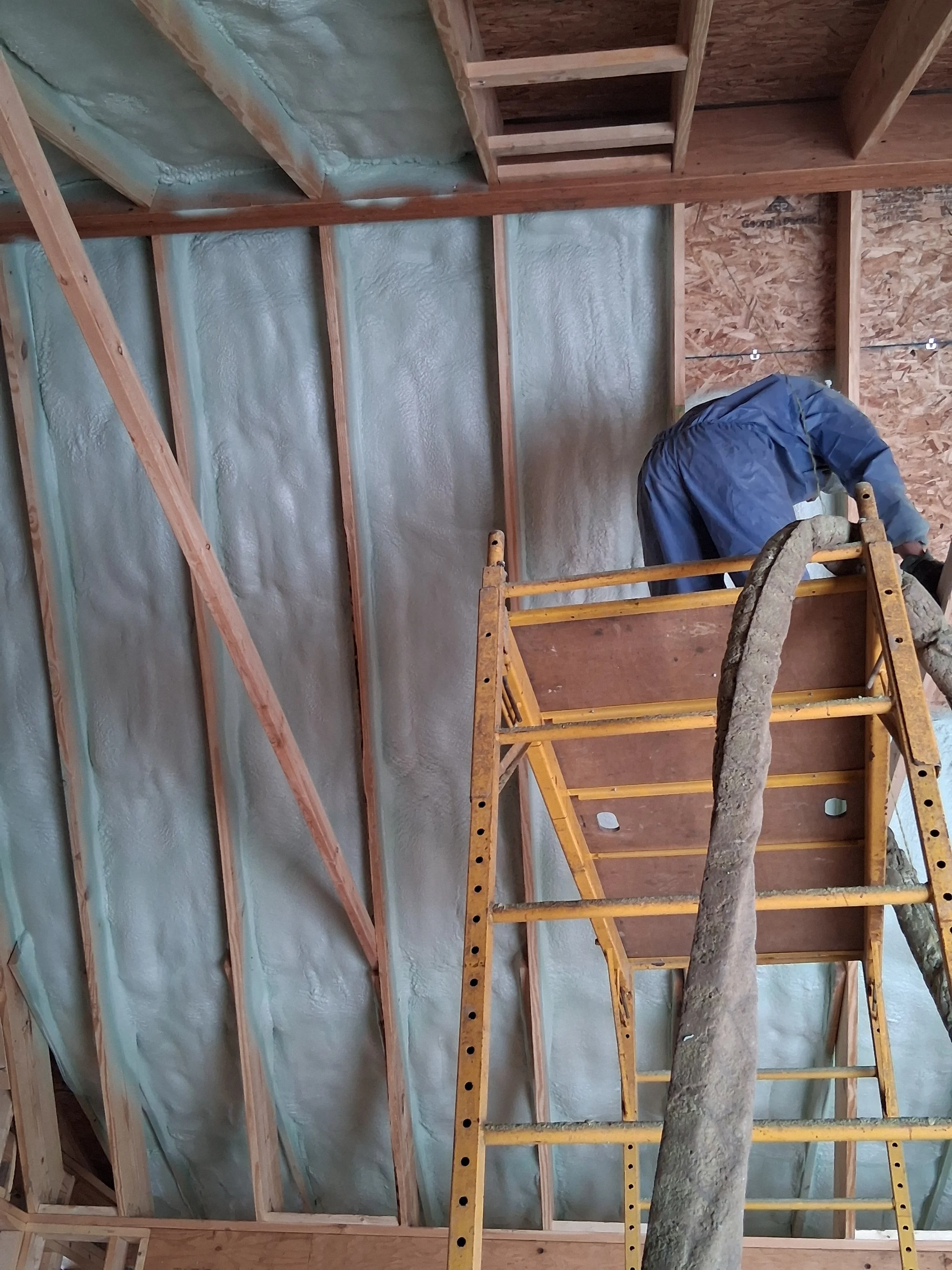 Construction worker in blue overalls installing insulation between wooden wall studs inside a building under construction.