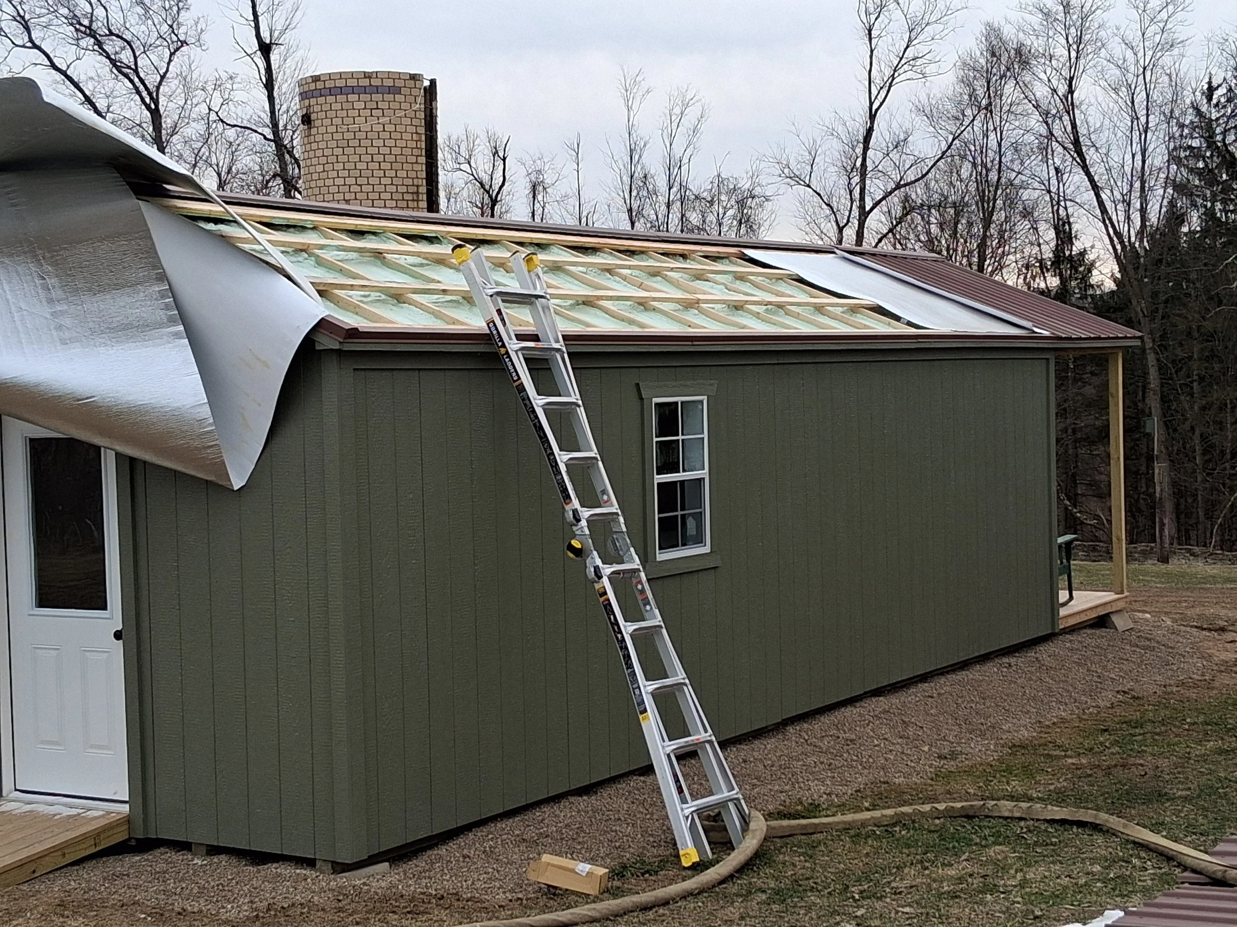 A house under construction with a green exterior, a ladder leaning against it, and a roof being replaced with new roofing materials. The yard is mostly dirt and gravel, with a hose on the ground.