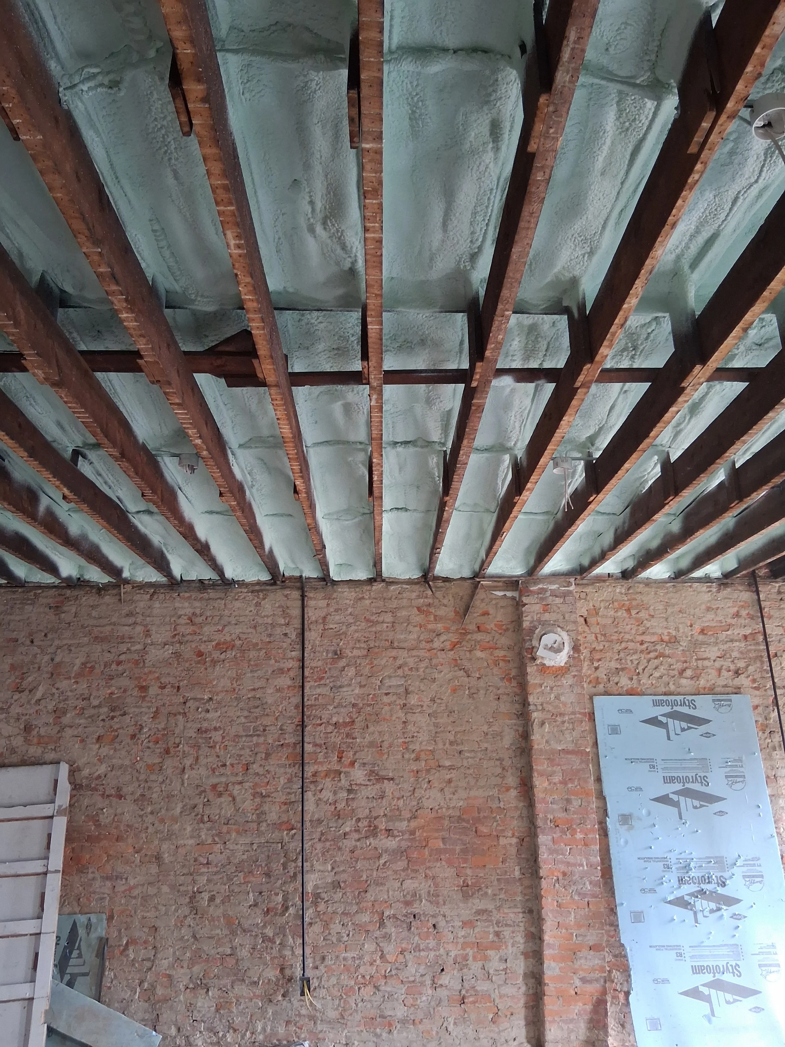 Construction site showing a brick wall, exposed wooden beams, and insulation material on the ceiling.