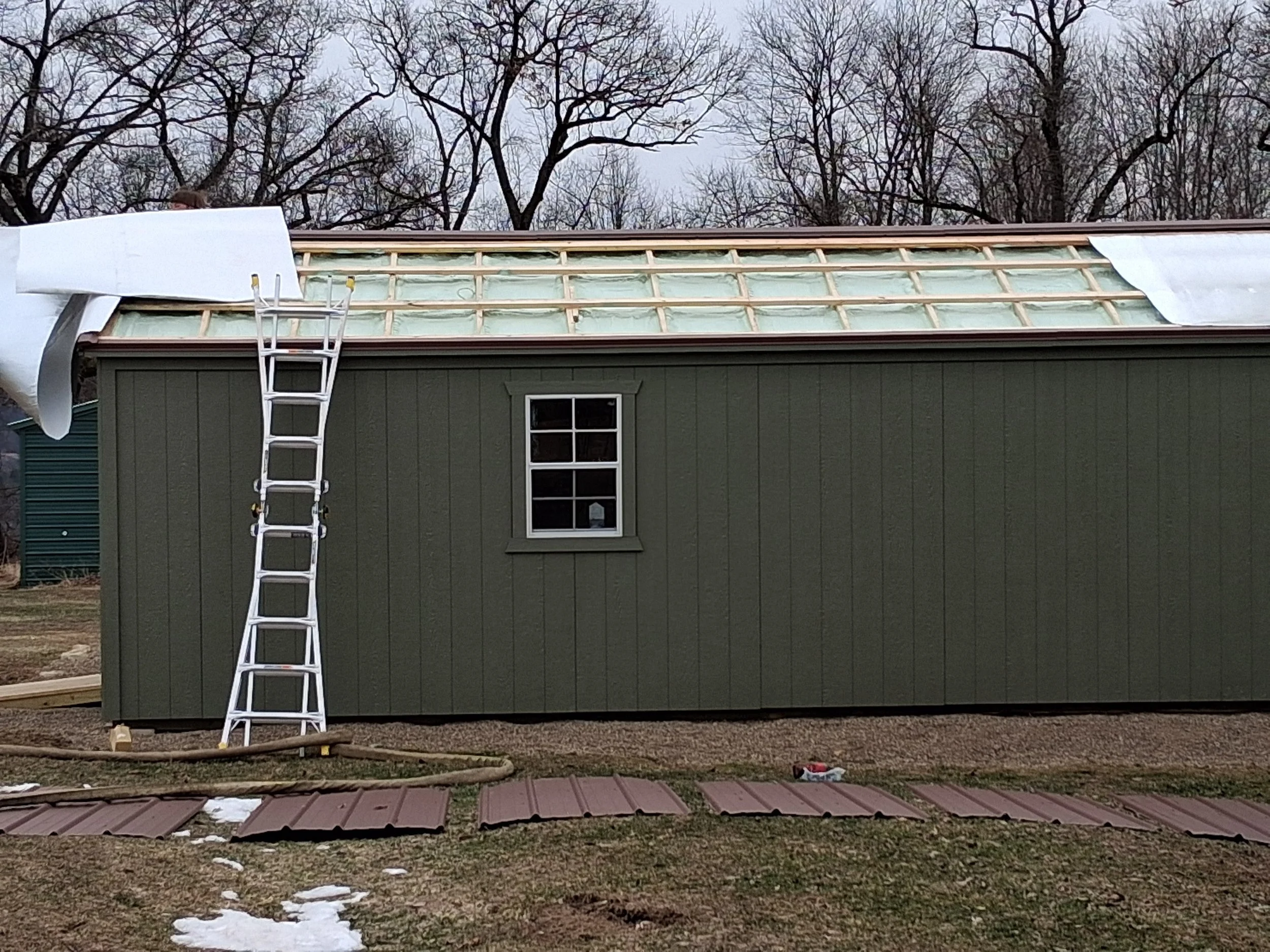 A house under construction with a roof partially covered in green sheathing and a ladder leaning against the side. Snow patches are on the ground, and there are construction materials around.