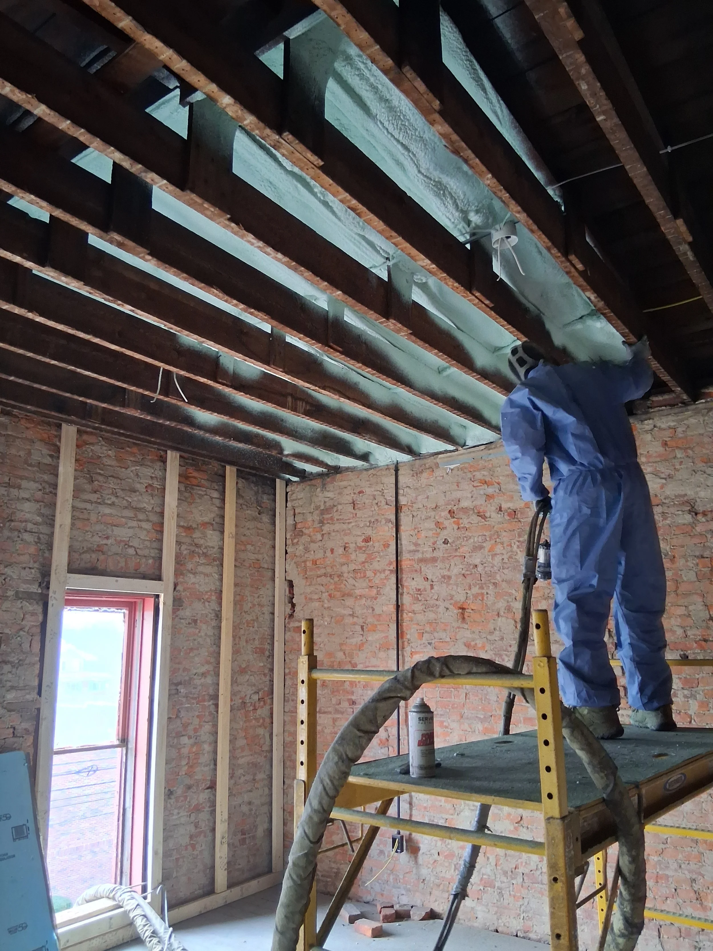A construction worker in protective gear sandblasting or working on the exposed ceiling of a room under renovation, with insulation material visible.