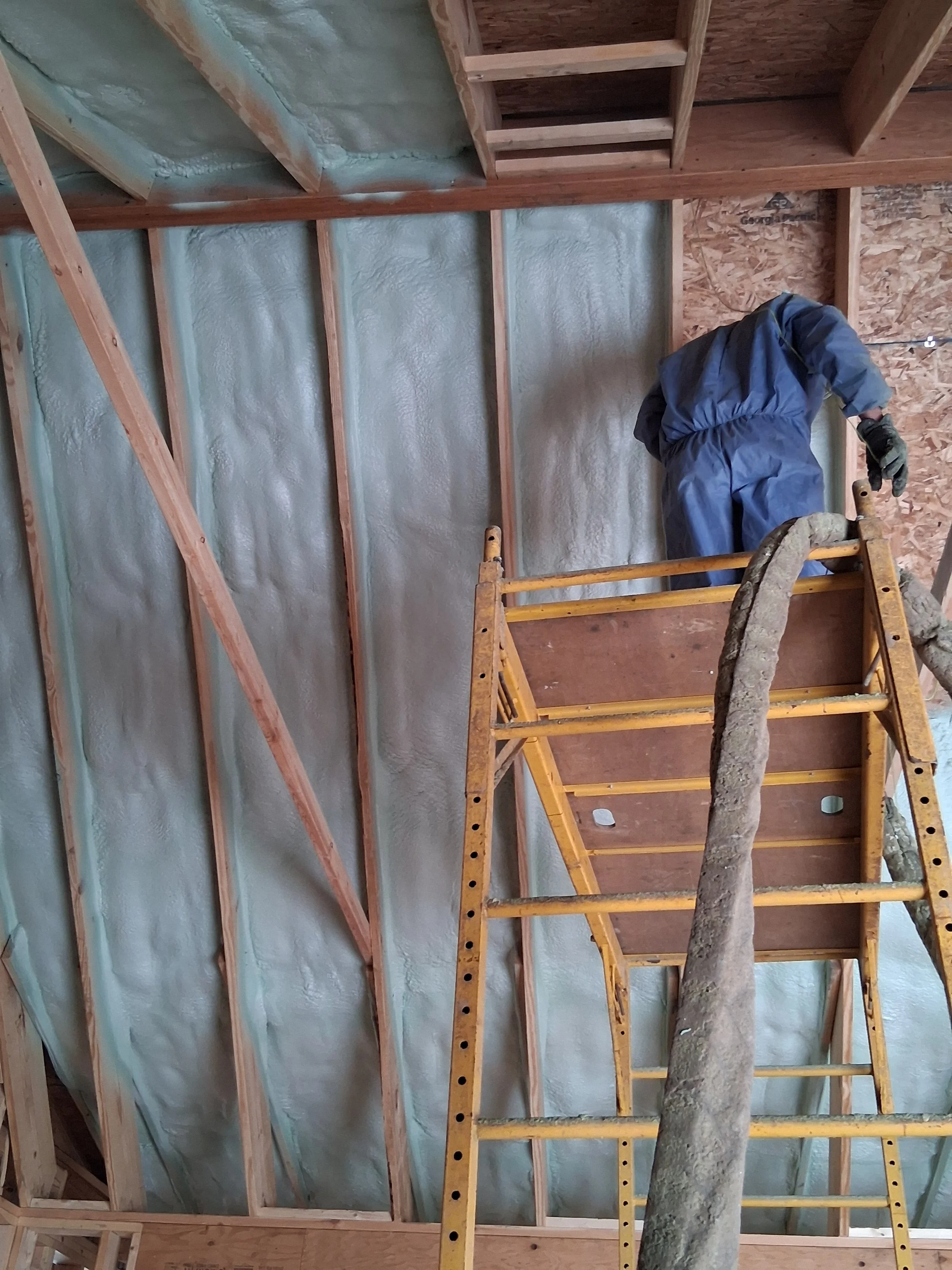 Construction worker in blue overalls standing on a scaffold working on insulation in a wooden framed room.