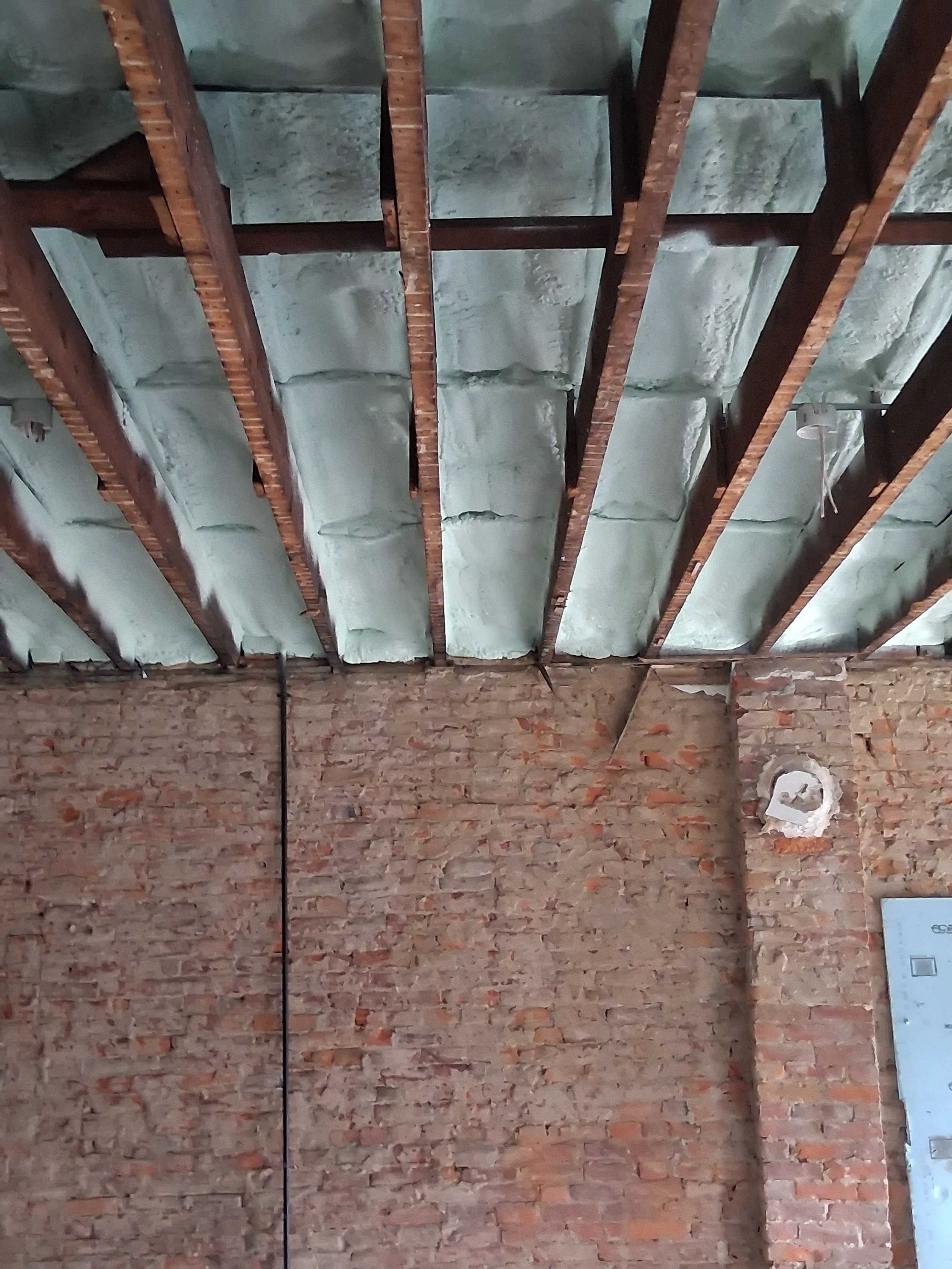 Interior of a building under construction with an exposed brick wall and a ceiling with insulation and wooden support beams.