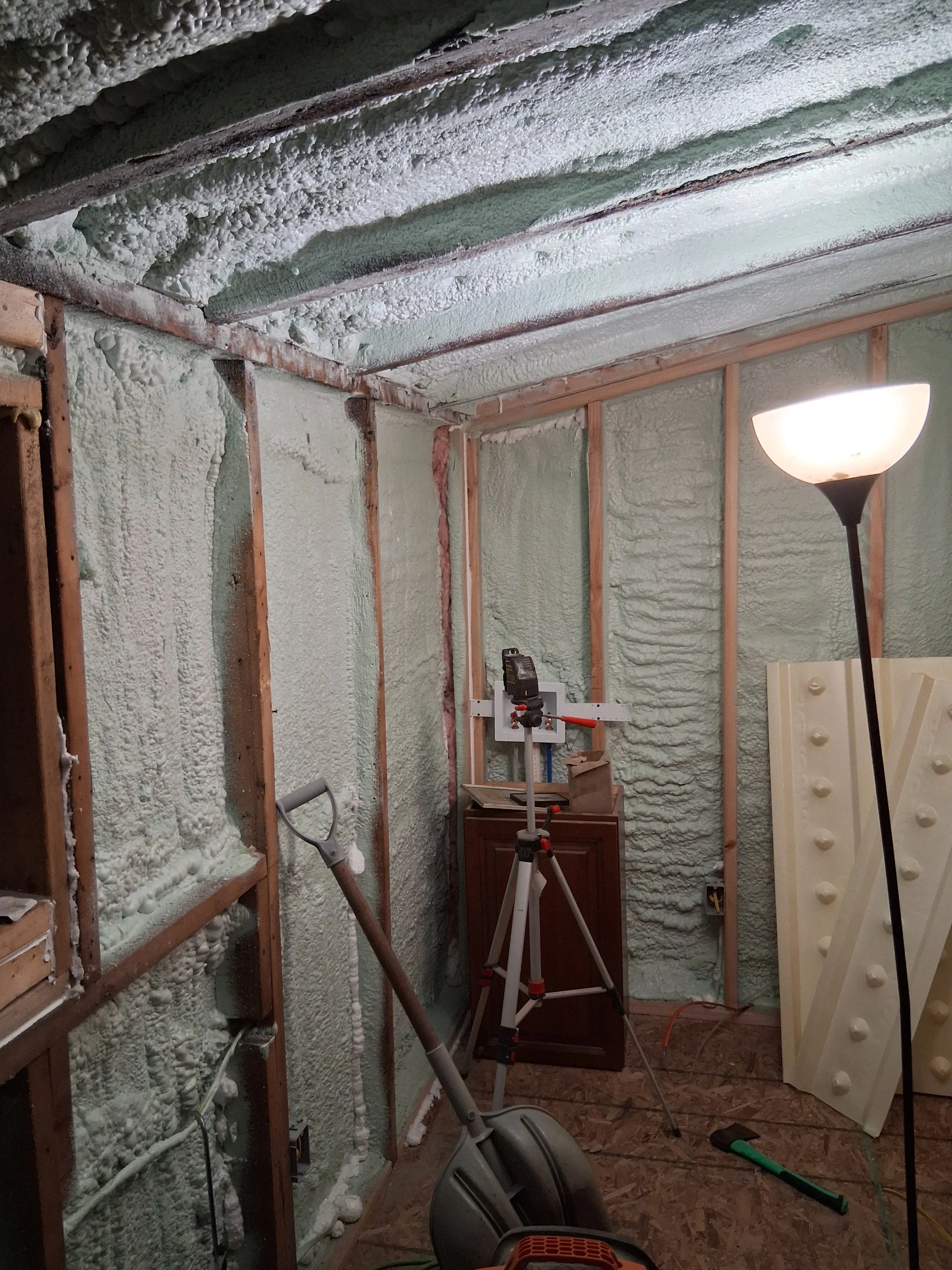 Room under construction with spray foam insulation on walls and ceiling, a floor lamp, a tripod with a tool, and various construction tools and materials.