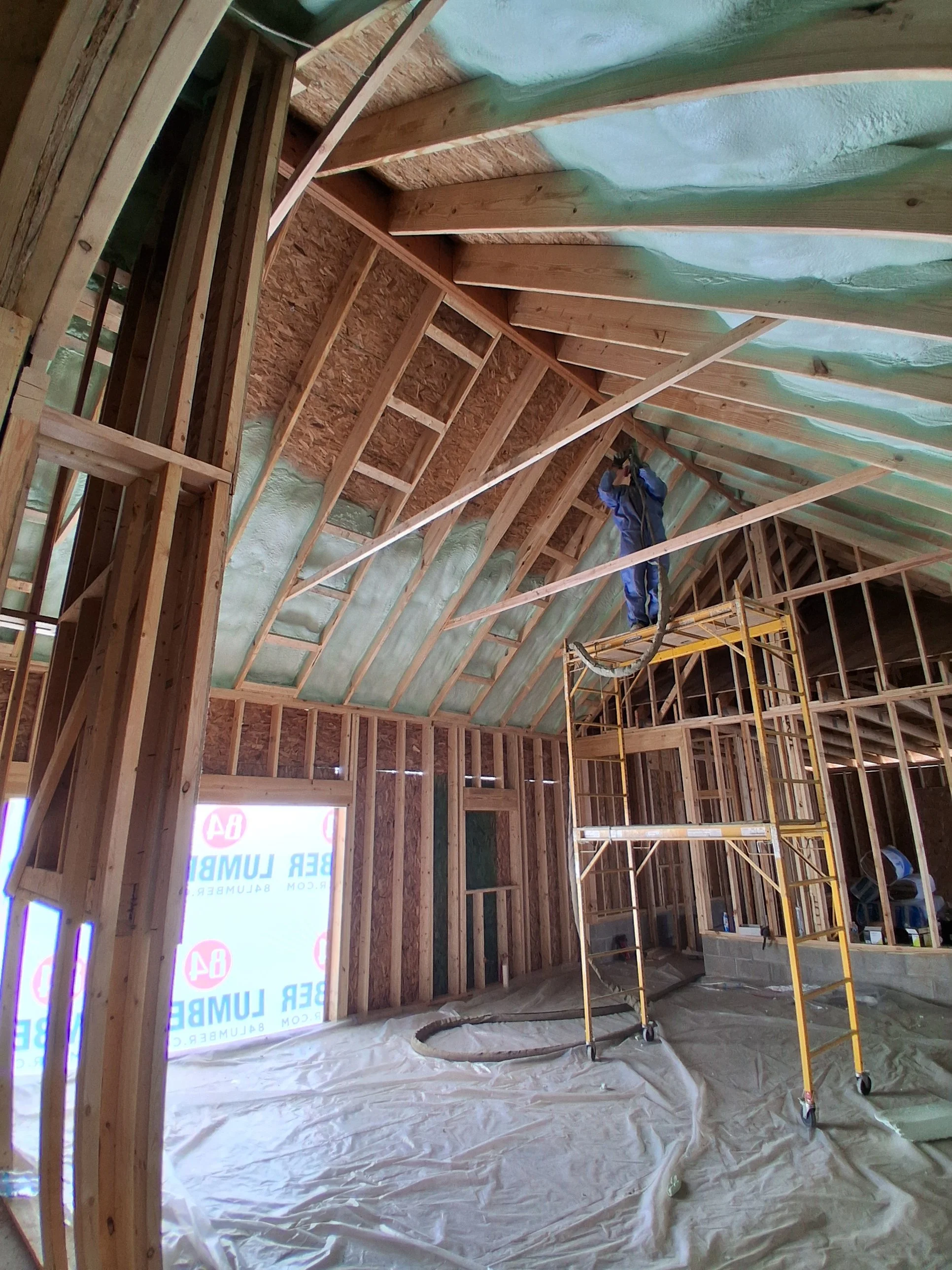 Construction workers installing insulation in the ceiling of a new house. The room is framed with wood, and a worker stands on scaffolding, working near the ceiling.