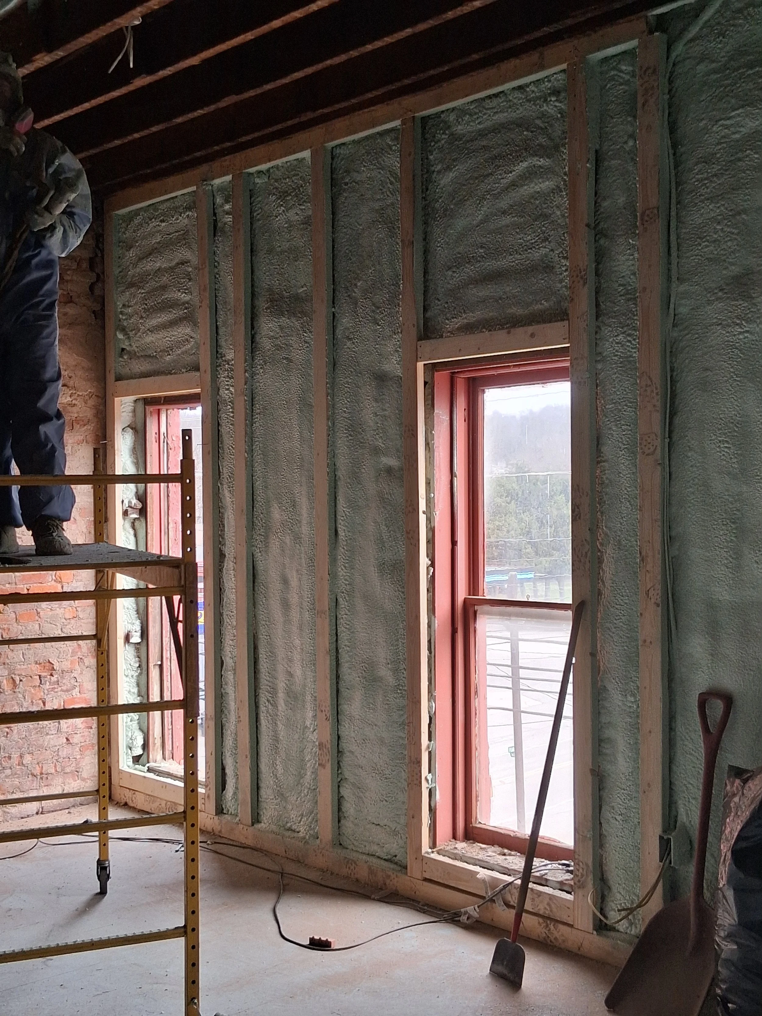 Indoor construction site with unfinished walls and insulation. There are two windows with red framing, a scaffold on the left side, and construction tools including a shovel and a pry bar. A worker is partially visible on the scaffold.