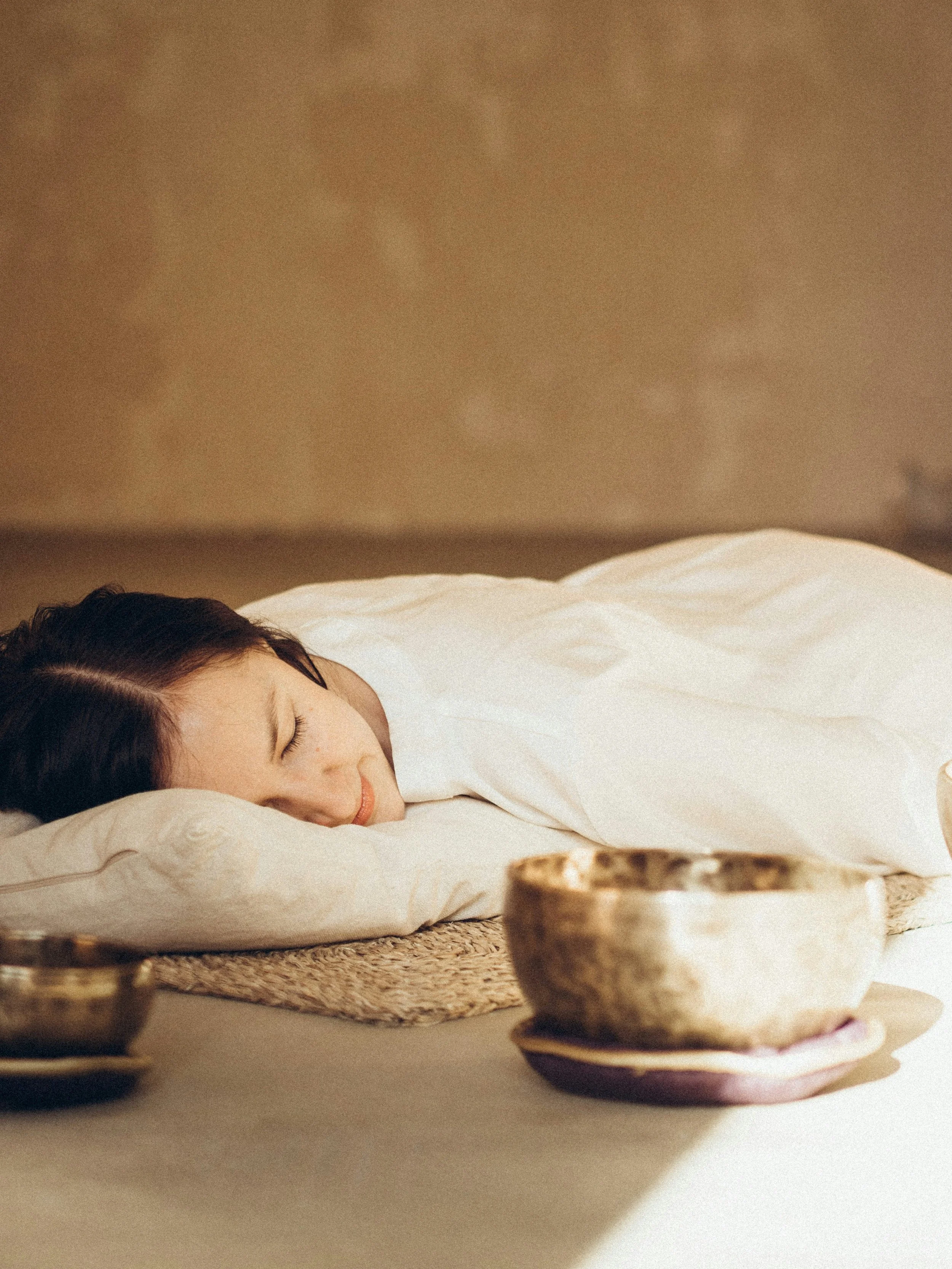 A woman sleeping peacefully on her side with her head resting on a pillow, covered with a white blanket, and surrounded by gold and ceramic bowls on a woven mat.