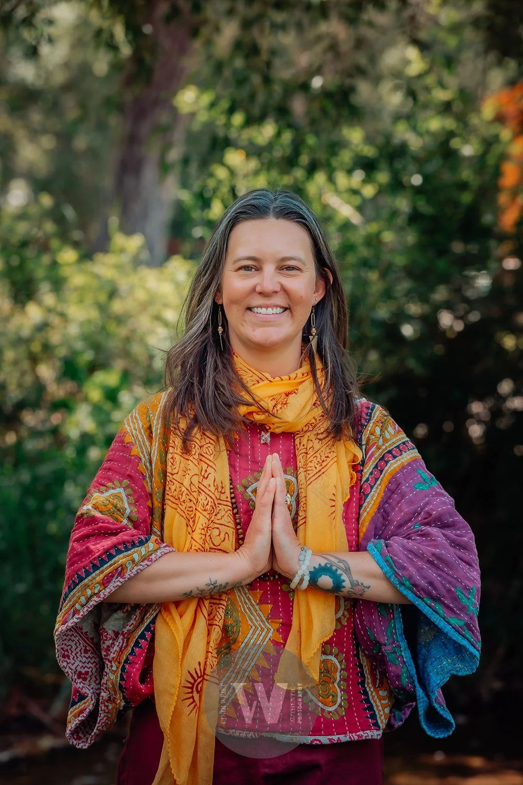 Lou Rae standing outdoors with green trees in the background, smiling, wearing a colorful patterned top and a yellow scarf, with her hands pressed together in a prayer pose.