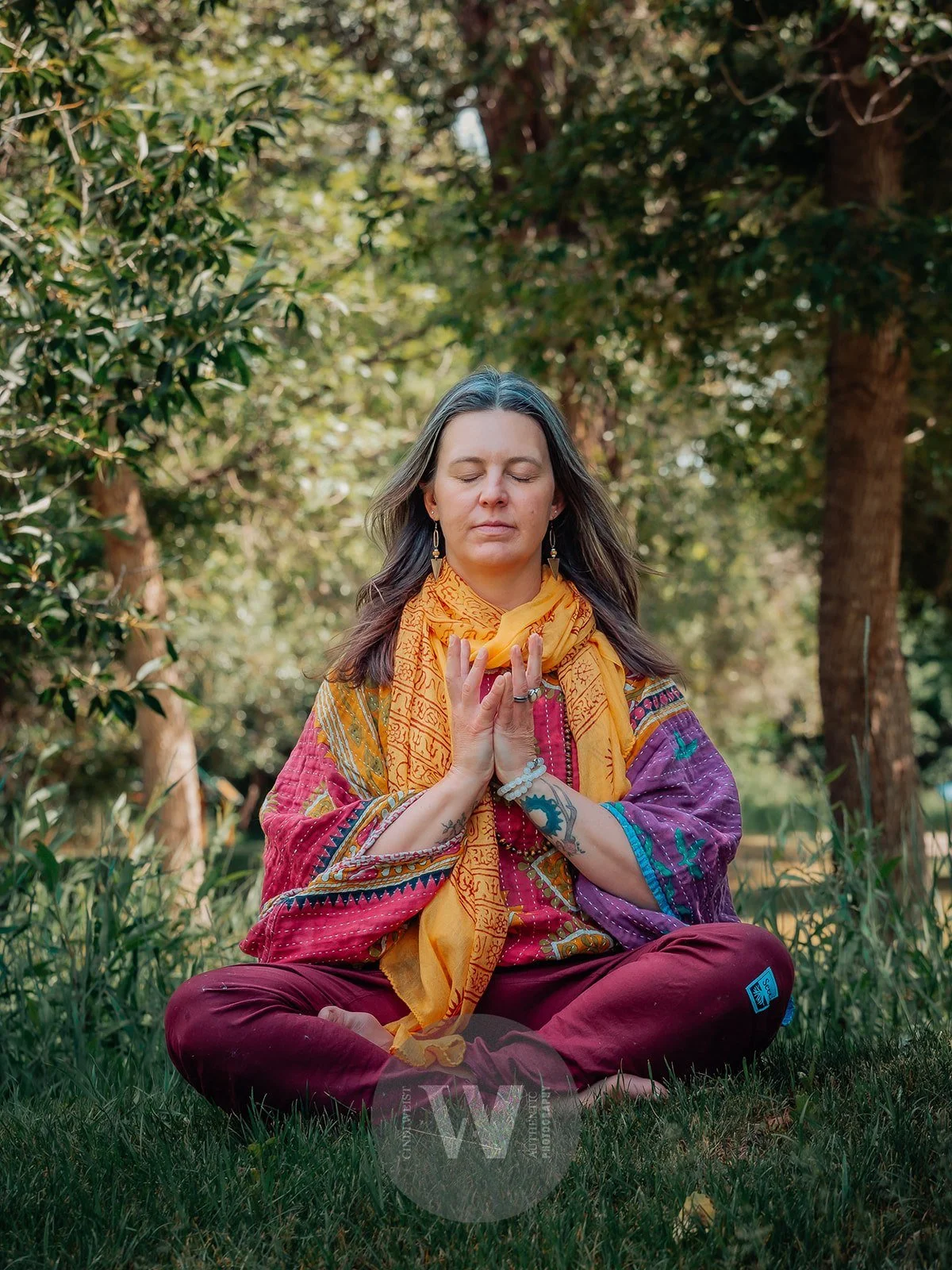 Lou Rae practicing yoga outdoors, seated cross-legged on grass by trees, with hands in prayer position, eyes closed, wearing colorful attire and jewelry.