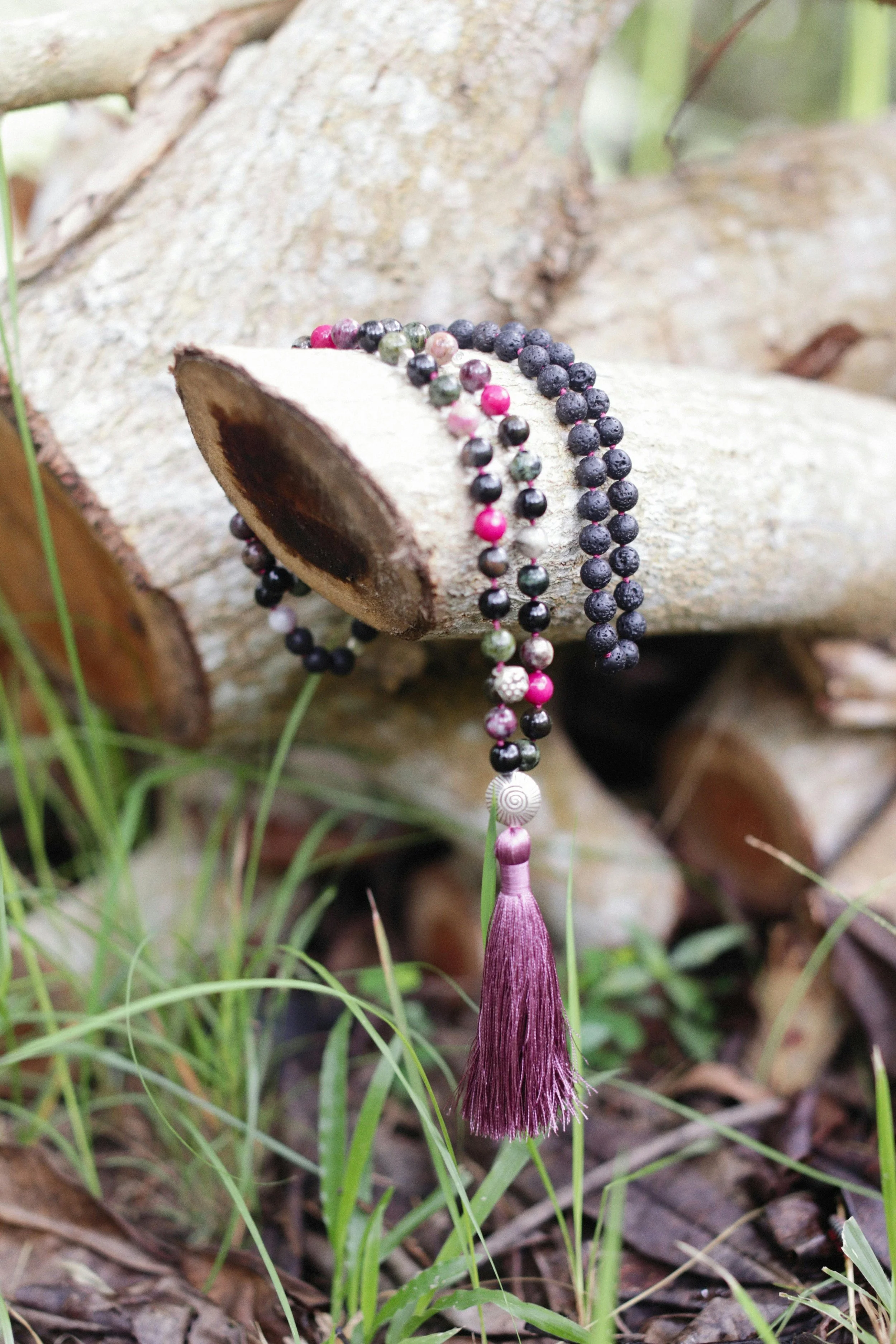 Close-up of a beaded necklace with a purple tassel hanging from a fallen tree trunk in a grassy area.