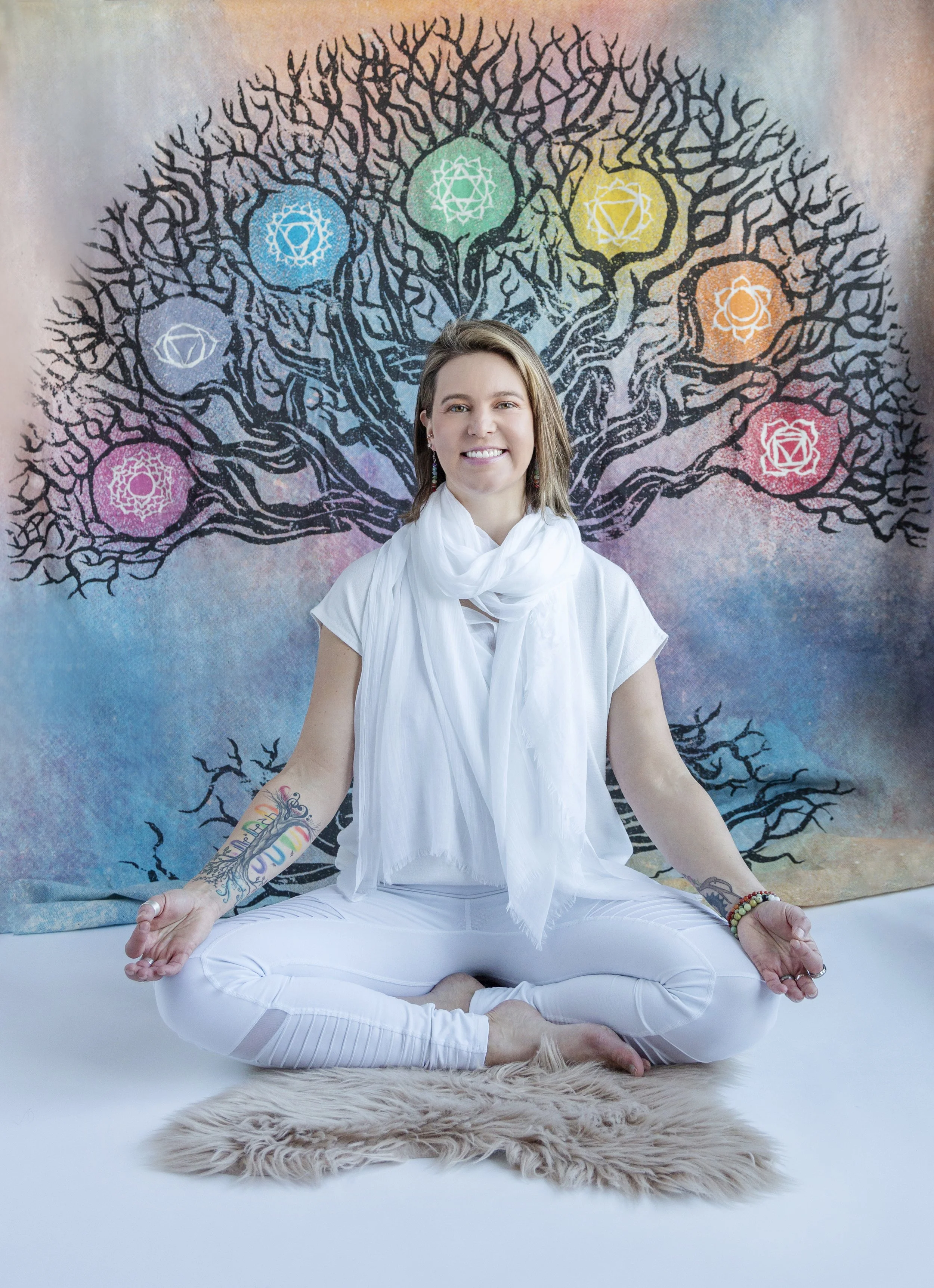 Lou Rae  practicing yoga in a seated meditation pose with legs crossed, hands resting on knees, wearing white clothing, and sitting on a furry rug in front of a colorful mural of a tree with chakra symbols.