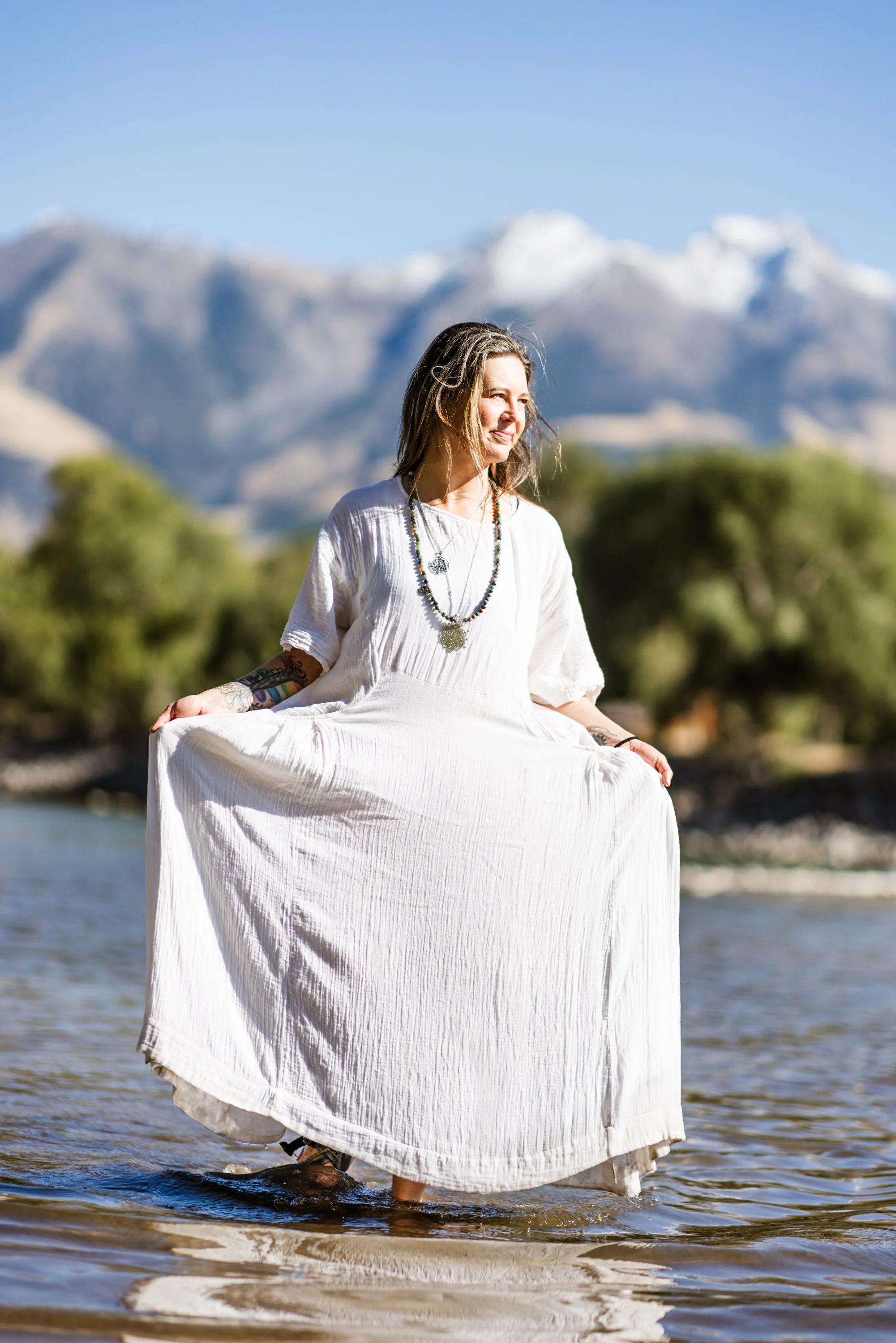 Lou Rae  in a white dress sitting in water, with mountains and trees in the background.