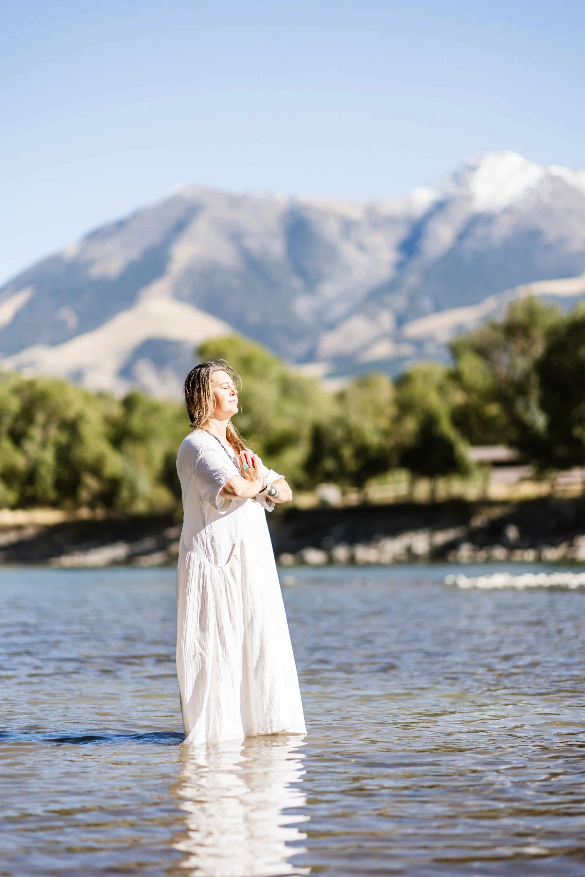 Lou Rae standing in a river with her eyes closed, wearing a white dress, with a mountain range and trees in the background, and the sky clear and blue.