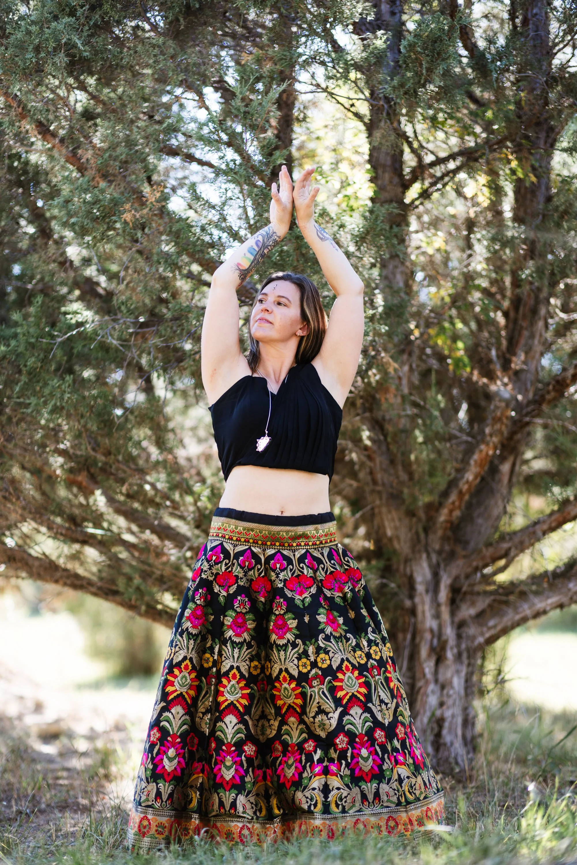 Lou Rae  standing outdoors in front of a large tree with green foliage, wearing a black top and a long embroidered skirt with vibrant floral patterns, and raising her arms above her head with her palms pressed together.