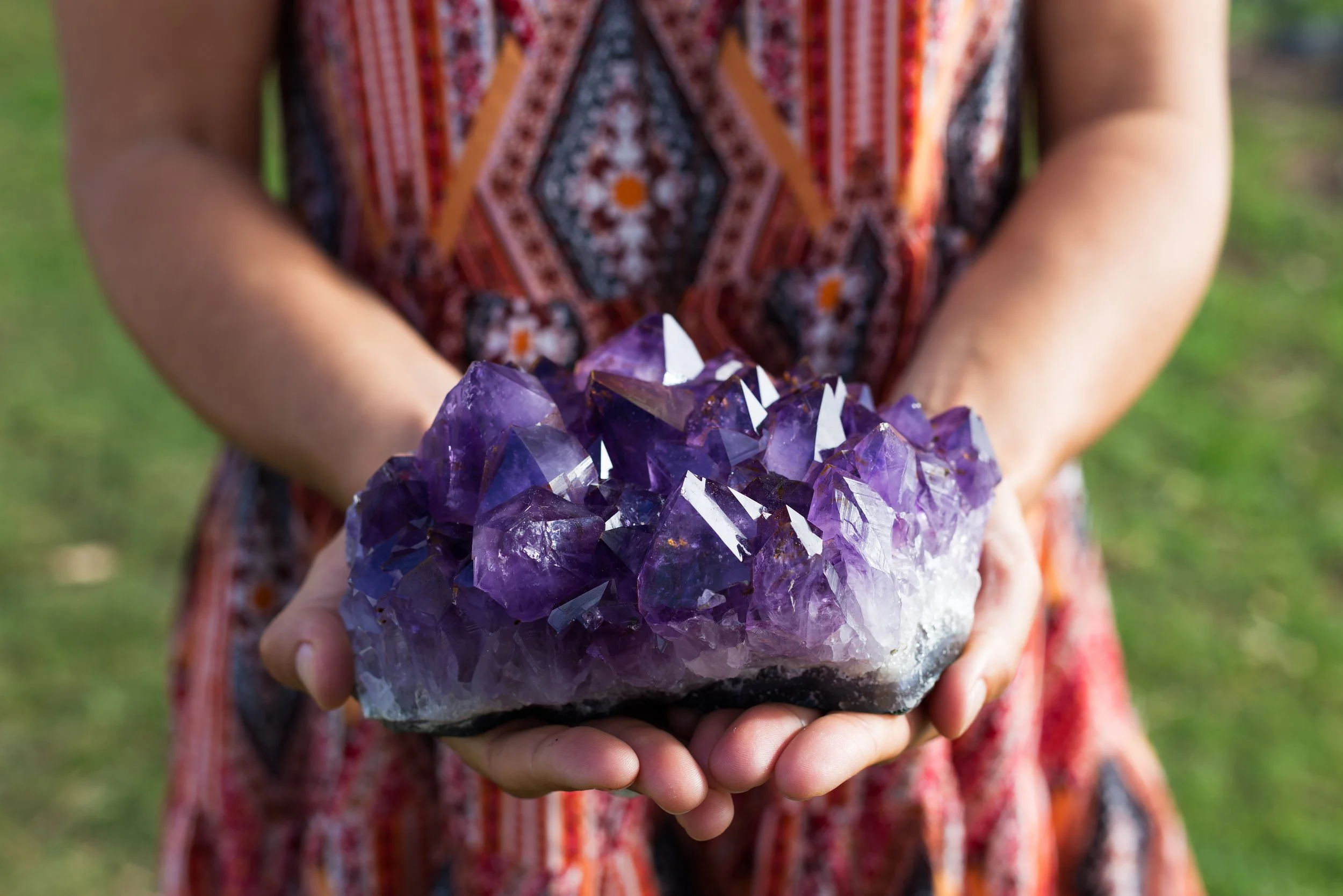 Person holding a large purple amethyst crystal cluster outdoors with a green background.