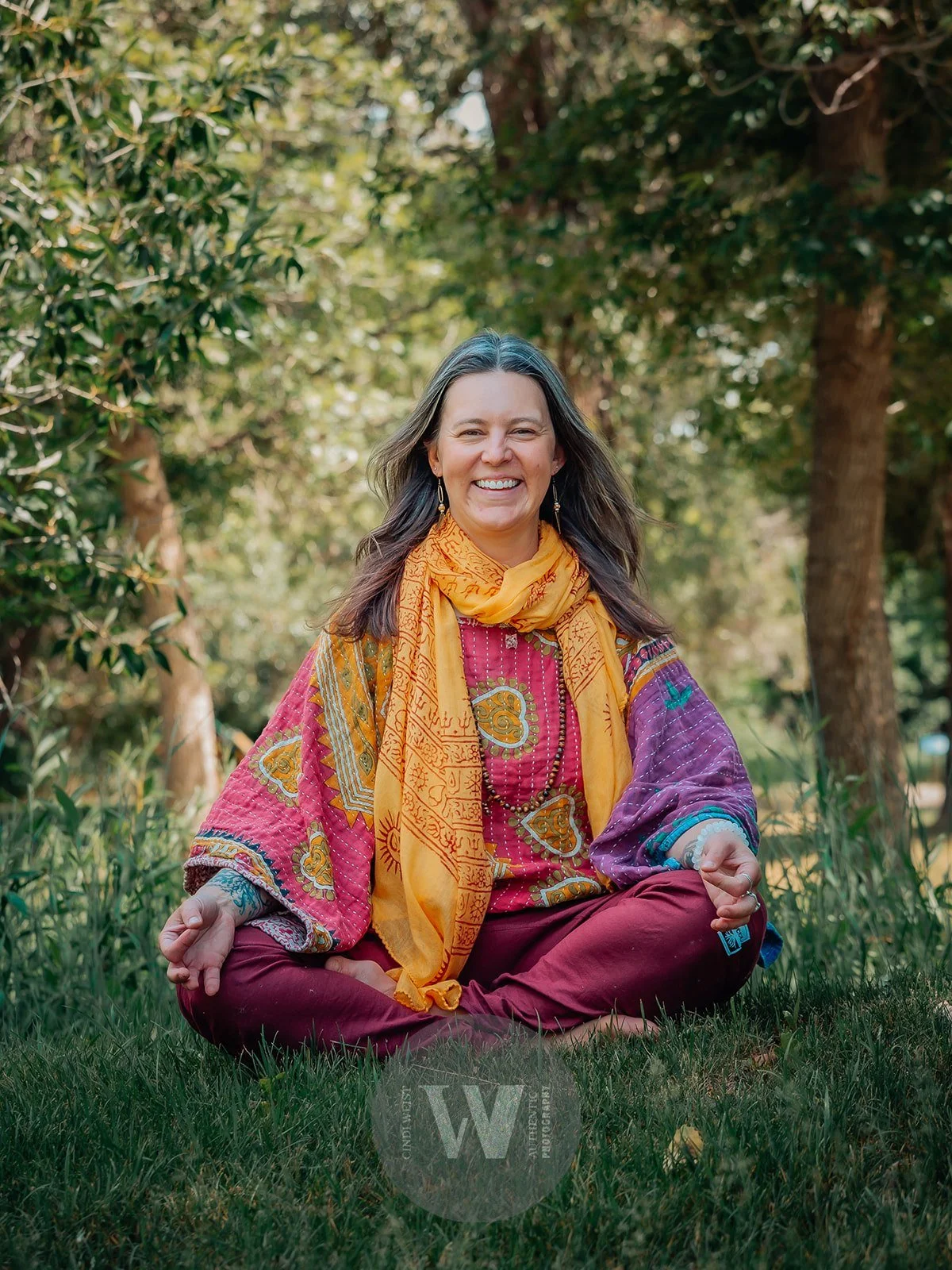 Lou Rae  sitting cross-legged outdoors on grass, smiling, with trees and greenery in the background. She is wearing a colorful, patterned blouse, a yellow scarf, and maroon pants.