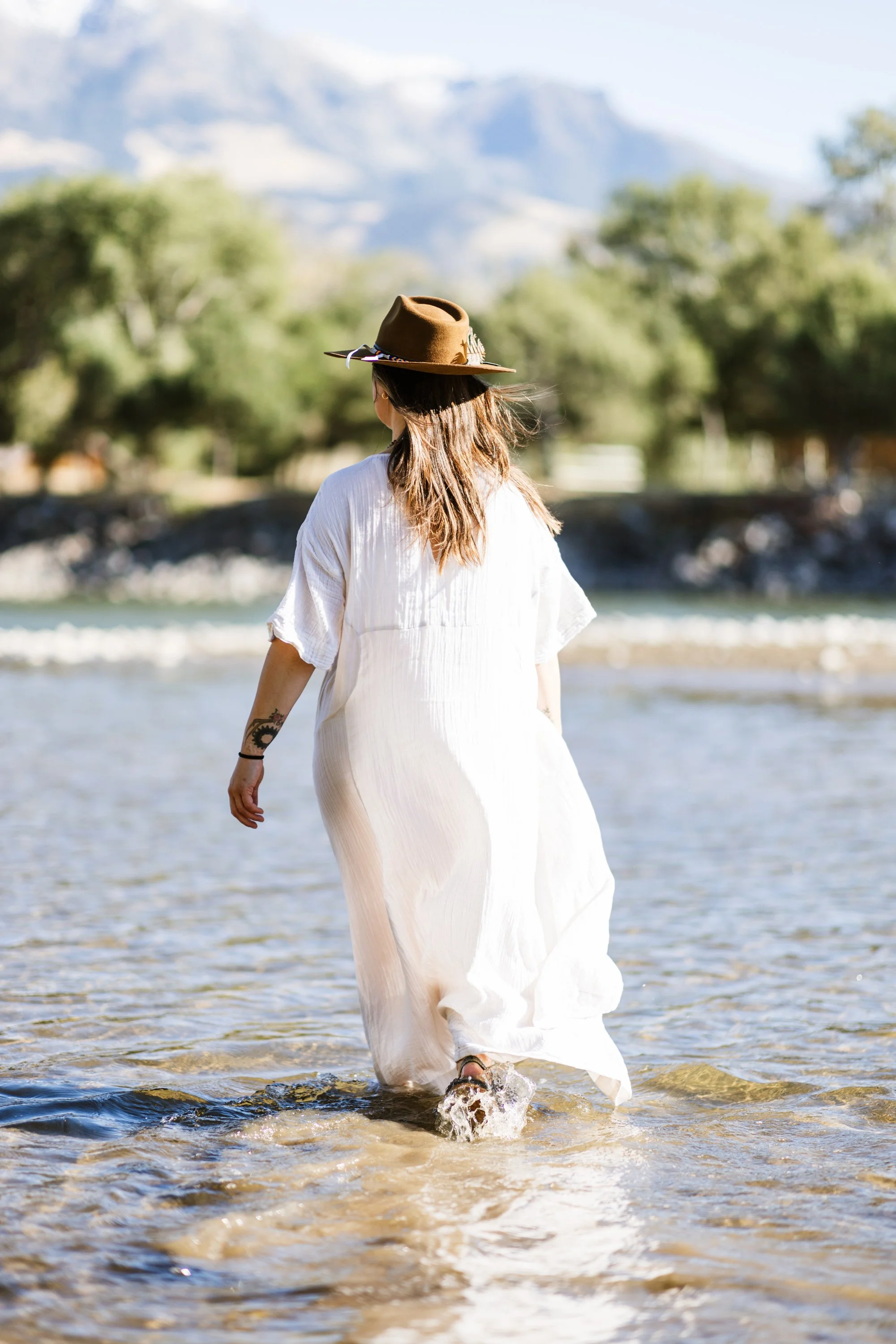 Lou Rae in a white dress and a brown wide-brimmed hat walking in shallow water at the beach, with greenery and mountains in the background.