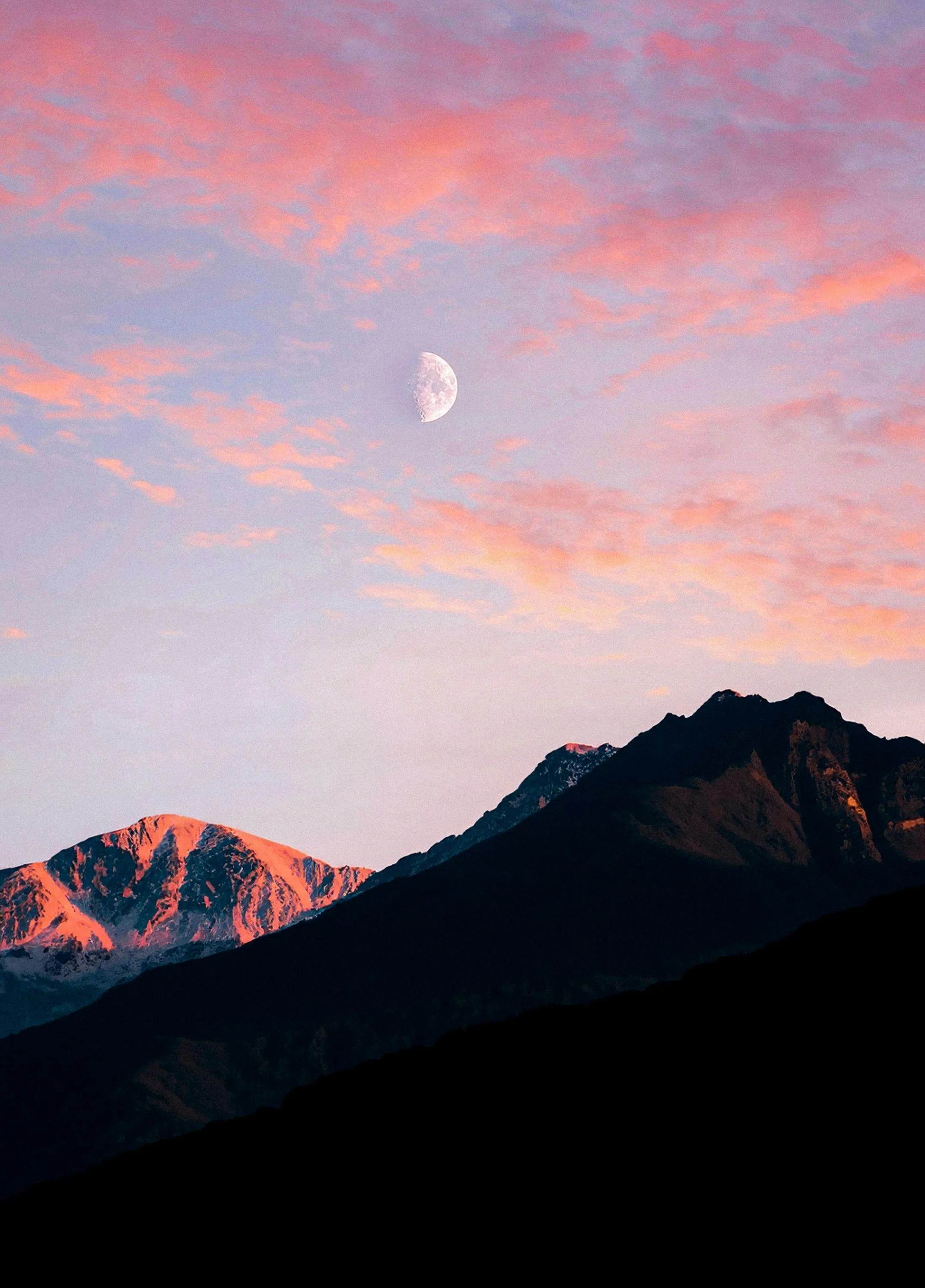 A mountain landscape during sunset with snow-capped peaks illuminated in pink and orange colors, dark silhouetted mountains in the foreground, and a pink and purple sky with the waxing gibbous moon visible.